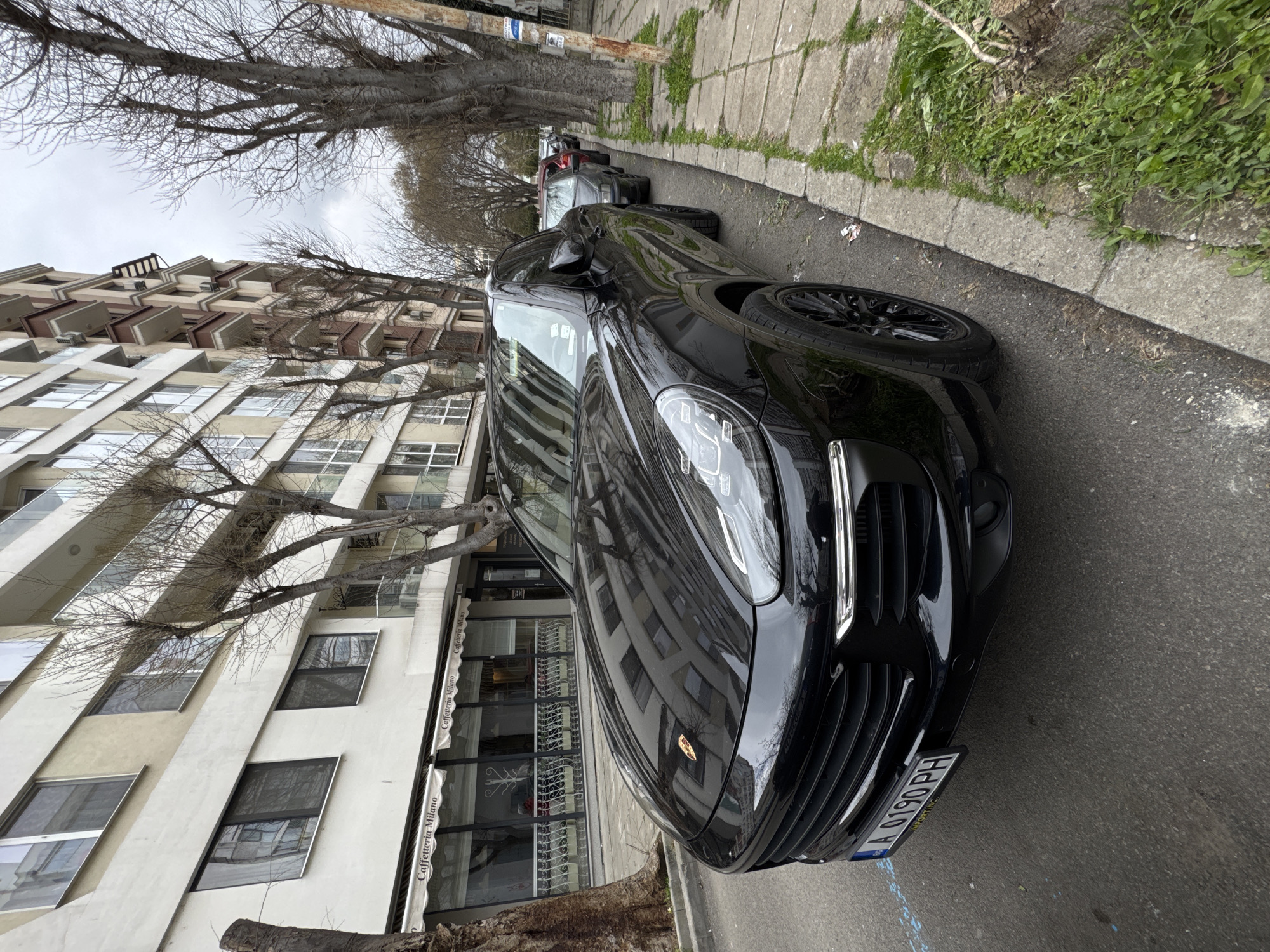 A black Porsche Cayenne is parked on a street next to a building in Burgas, Bulgaria. The building has a "Caffetteria Milano" sign. Other vehicles are parked further down the street. Bare trees line the sidewalk. The weather appears overcast. The Porsche has a license plate that reads "A 0190 PH".