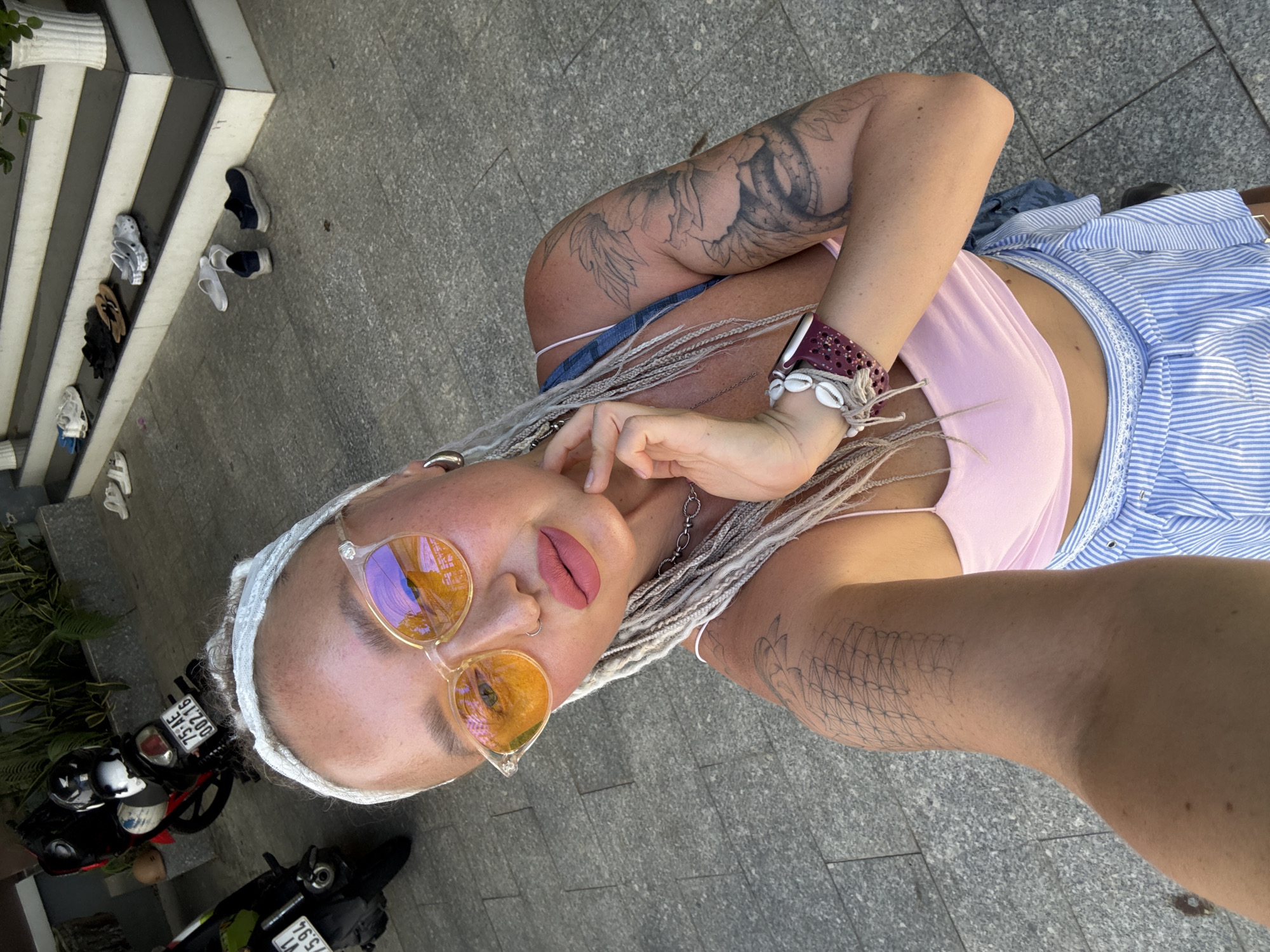 This is a portrait-style, top-down selfie of a person outdoors in what appears to be a city environment, possibly Nha Trang, Vietnam. The subject has light skin, grey braided hair, and is wearing a pink tank top and a blue and white striped garment that looks like a skirt or dress. They are also wearing large, translucent orange-tinted sunglasses, a white headband, and a bracelet adorned with shells. Tattoos are visible on their left arm. The background shows a gray, tiled ground and some greenery, as well as several motorcycles parked nearby. The light suggests it is daytime, likely sunny. The subject is touching their face with their fingers, and their expression, with the sunglasses and makeup, is somewhat alluring. There are license plates on the motorcycles with visible text, including "75-AE 007.16" and "75-A1 75.94". Several pairs of shoes are also visible in the background on some steps.