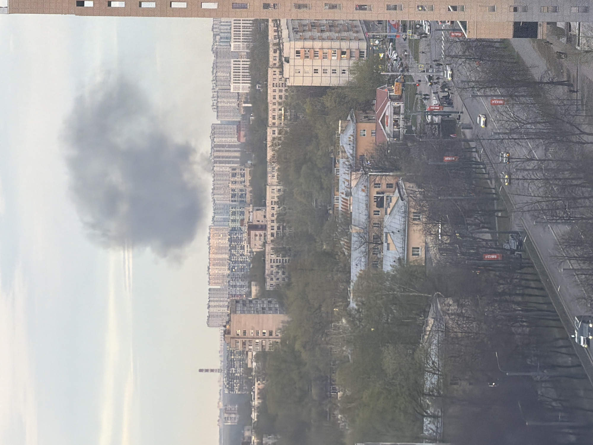 The image shows an urban landscape, likely in Russia. There are several rows of tall apartment buildings stretching into the distance, interspersed with green trees. A road runs parallel to the buildings, with cars and buses visible. There are also signs, possibly street signs, along the roadside. In the upper left of the image is a large grey cloud. The photo appears to be taken from a high vantage point, possibly from another building.
