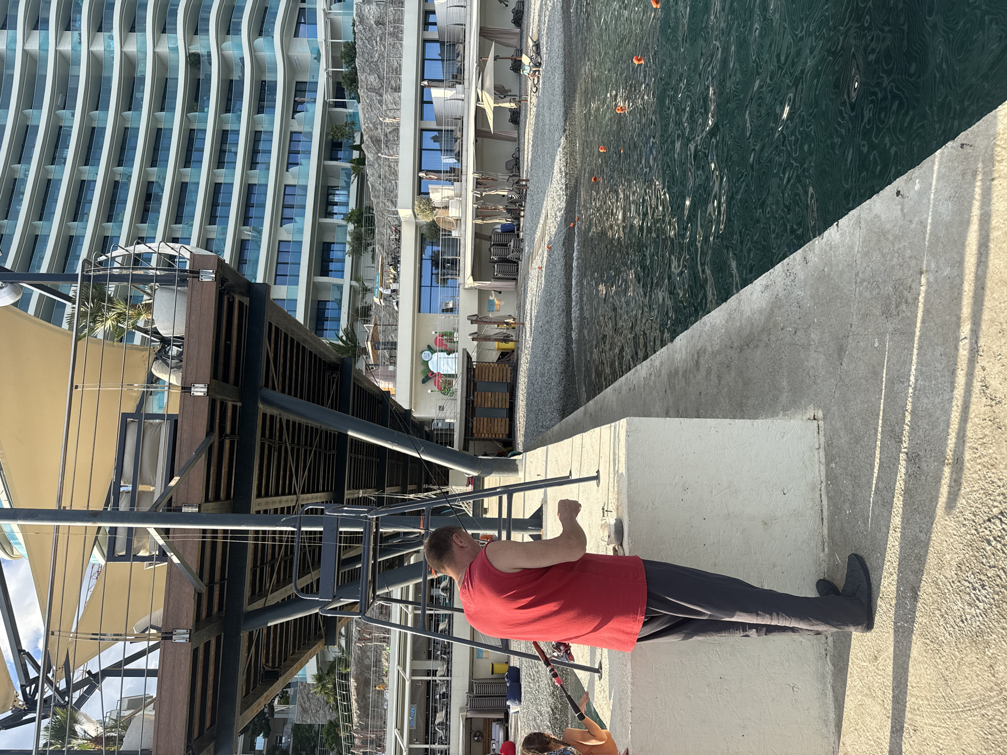 In a coastal scene set in Yalta, Ukraine, a man in a red tank top and dark pants stands on a concrete pier, facing away from the camera. He appears to be fishing, holding a fishing rod with a reel. The pier overlooks a body of water with gentle ripples and orange buoys scattered across its surface. In the background, a modern, multi-story building with a distinctive wavy facade rises, featuring numerous windows and balconies. Adjacent to the building is a beach area with rows of stacked chairs and umbrellas, suggesting a resort or hotel. The sunlight casts long shadows on the pier, indicating it is likely late afternoon. The overall atmosphere seems calm and relaxed, with the man engaged in a leisurely activity.