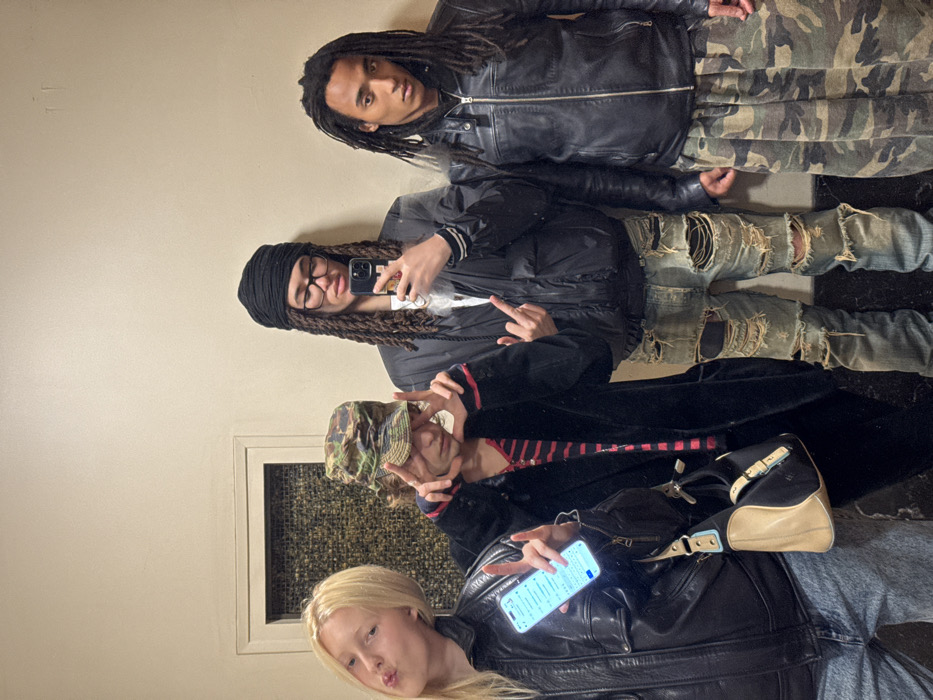 The image depicts an indoor mirror selfie of four individuals within an unidentified building in Paris, France. The background consists of a plain, light-beige wall, with a framed rectangular panel of textured, dark material visible on the far left.
From left to right:
1. A young blonde-haired individual with fair skin, wearing a black leather-look jacket and light blue jeans. They are pouting their lips and holding a brightly illuminated smartphone, displaying text, in their right hand.
2. An individual wearing a camouflage bucket hat that obscures most of their face, a long black coat over a red and black striped shirt, and dark trousers. They are making a peace sign (V-sign) with both hands. A black and tan shoulder bag is slung over their left shoulder.
3. An individual with dark skin, wearing a black fabric head wrap, eyeglasses, a black puffer jacket over a white top, and heavily ripped light blue jeans. This individual holds a dark-colored smartphone, positioned to capture the mirror selfie, in their right hand. Their left hand is raised, displaying a middle finger gesture.
4. A young male with dark skin and long dreadlocks, dressed in a black leather jacket over a black top, and loose-fitting camouflage pattern trousers. He stands with a neutral expression, looking directly at the camera reflection.
All subjects are posing for the camera reflected in the mirror.