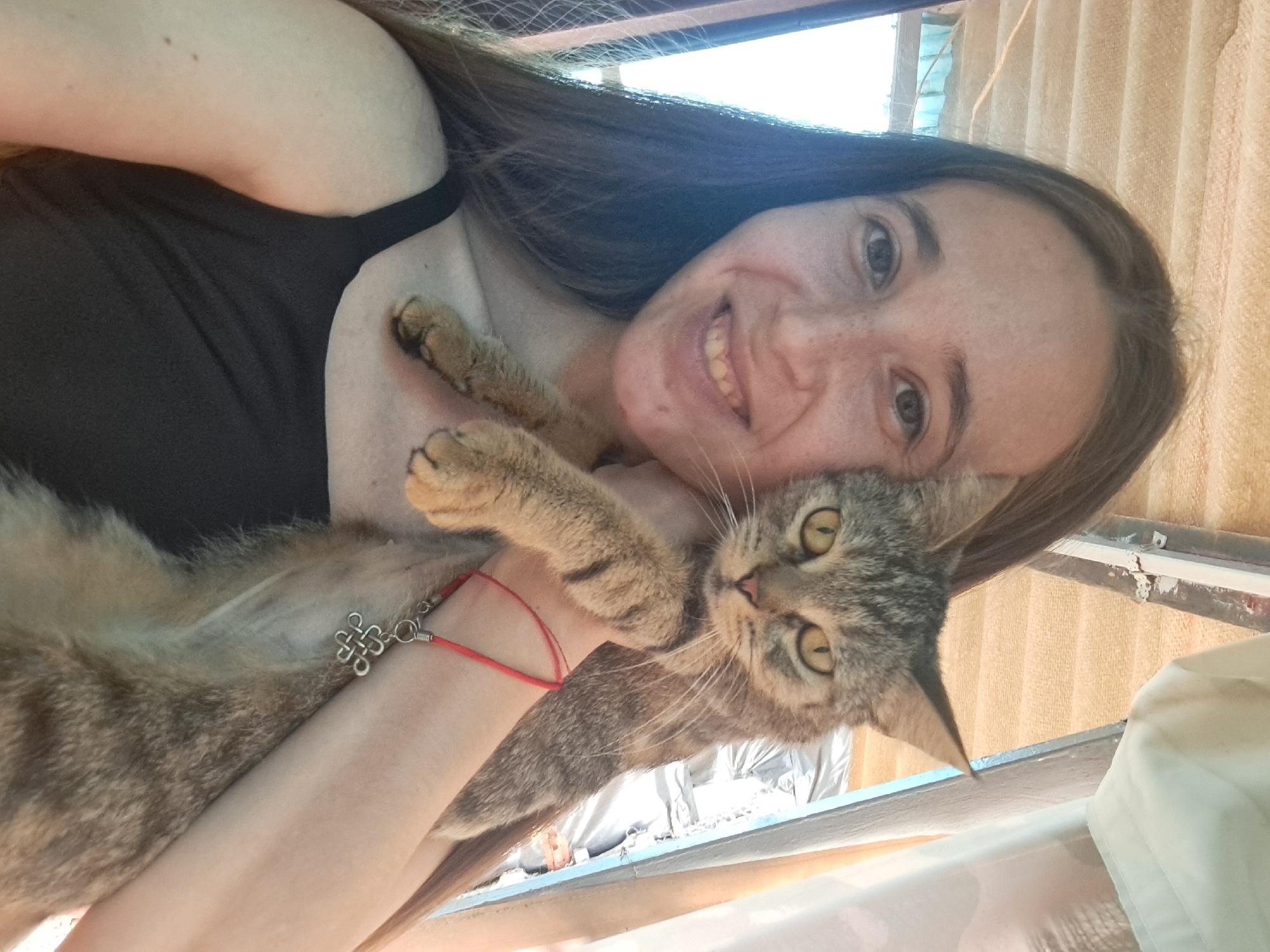 A young woman with long brown hair, freckles, and a black tank top is taking a selfie while holding a tabby cat. The cat is held in front of her chest, with its paws resting on her arm. A red string bracelet with a silver charm is visible on the woman's wrist. The background suggests an indoor or covered outdoor space with corrugated roofing and some draped fabric. There is no visible indication of the city of Vypasne, Ukraine.