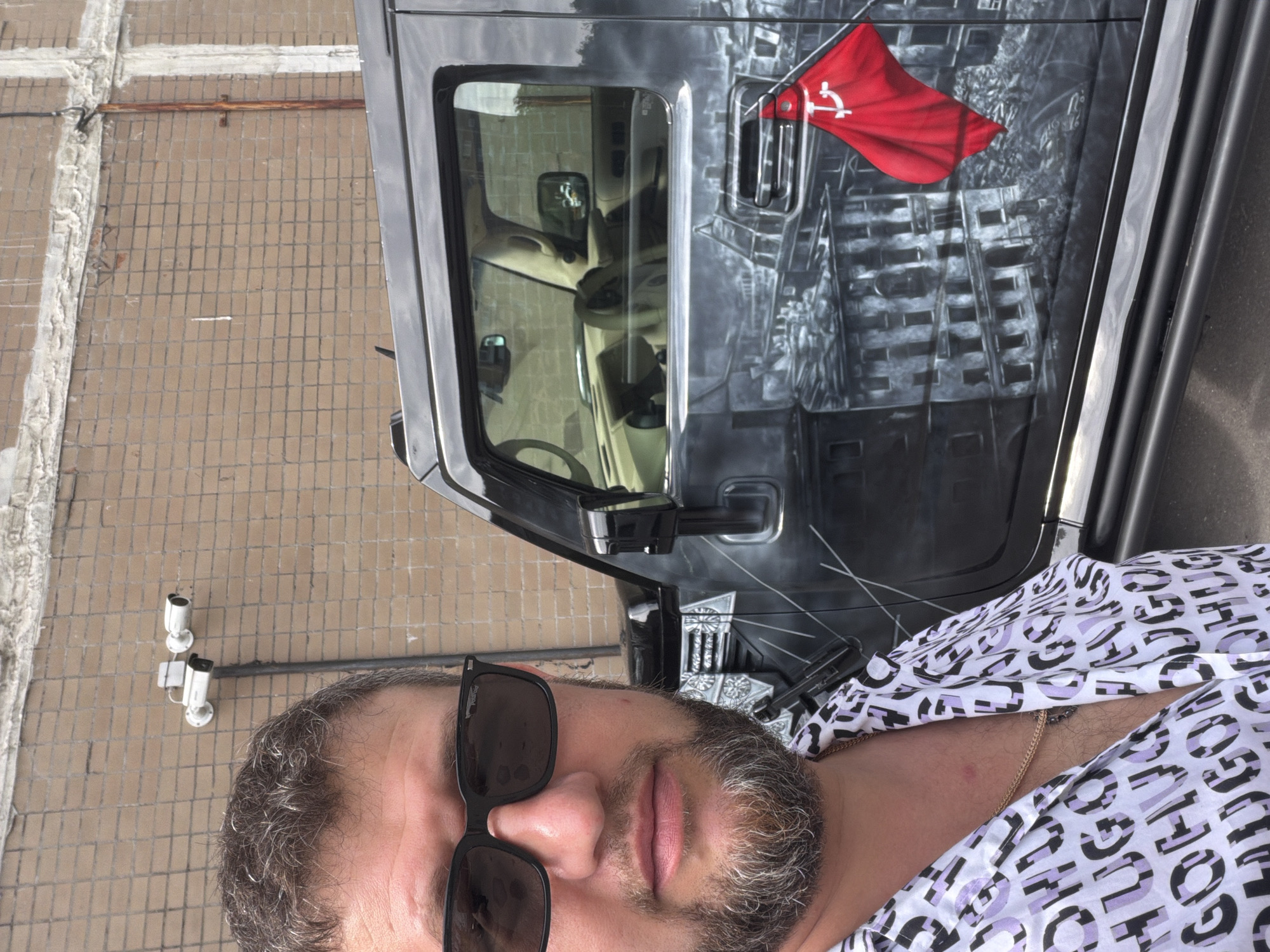 A man with short, graying hair, a beard, and sunglasses stands in front of a black SUV. He is wearing a white shirt with a repeating black and purple pattern of the word "HUGO" and a gold chain. The SUV has a mural painted on its side depicting a cityscape with a red flag featuring a hammer and sickle. The background is a brick wall with security cameras mounted on it. The scene appears to be outdoors in an urban setting.