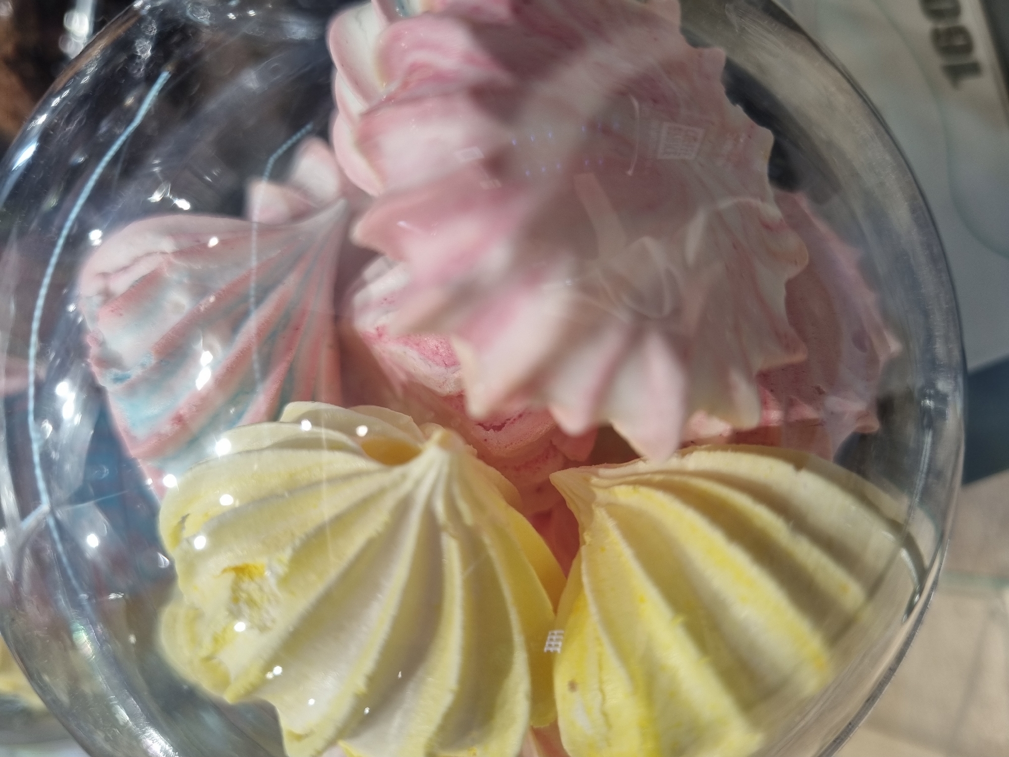 An overhead shot shows a glass bowl containing several meringue cookies in Sochi, Russia. The meringues are piped into a swirled, rosette shape. Two are bright yellow, and the others are white and pink. The glass bowl is slightly blurred by condensation and has a thick, rounded rim. The background is indiscernible due to the shallow depth of field.