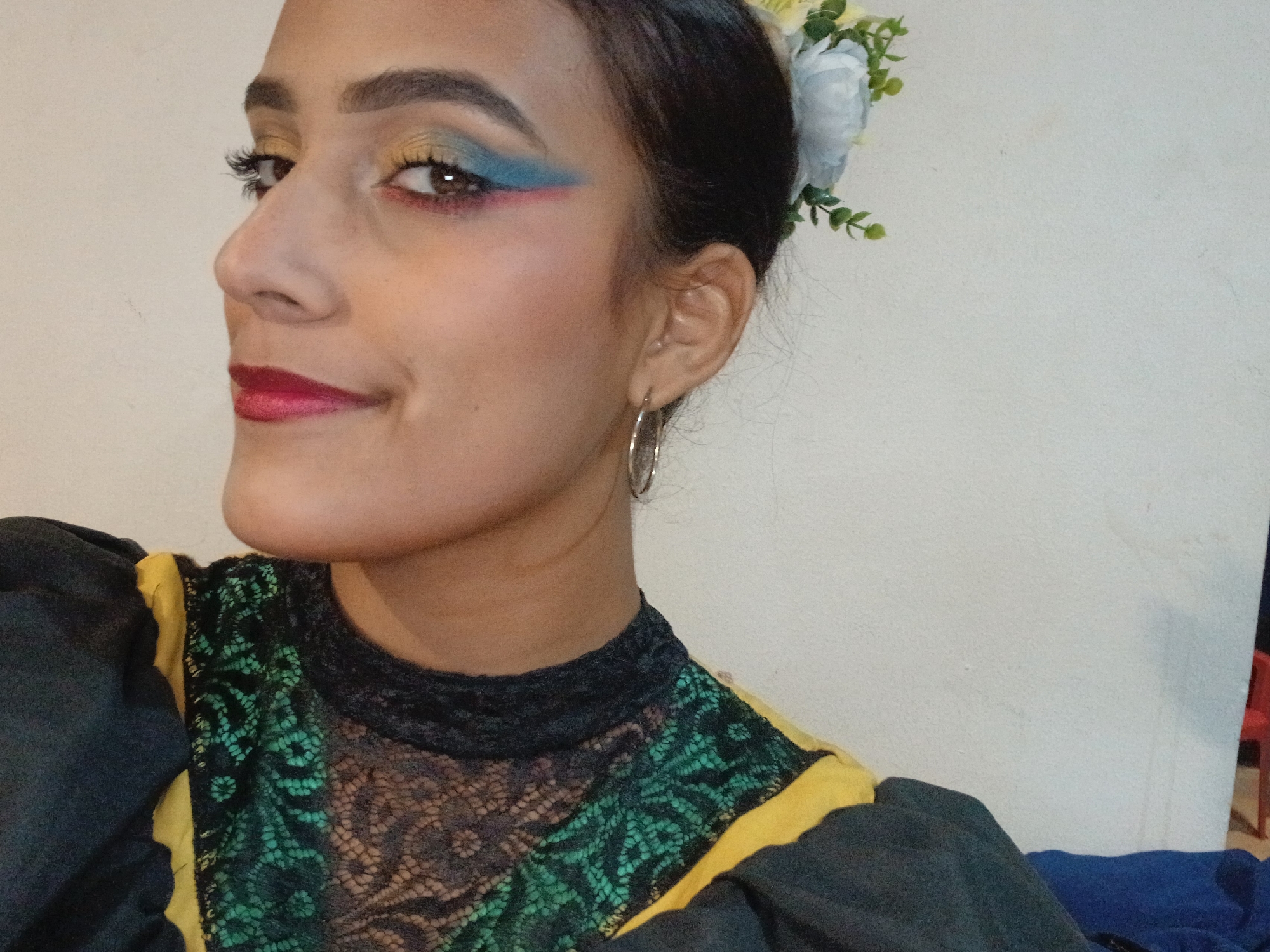 A woman is pictured in a close-up, slightly angled, head and shoulders shot. She is fair-skinned with dark eyebrows and brown eyes, wearing vibrant makeup featuring yellow, blue, and red eyeshadow, and red lipstick. A floral arrangement is in her hair near her ear, and she is wearing hoop earrings. Her top is a combination of black lace over a green and yellow fabric, with black sleeves. The background is a simple, light-colored wall. The image lacks definitive landmarks, so it is not possible to verify the location.