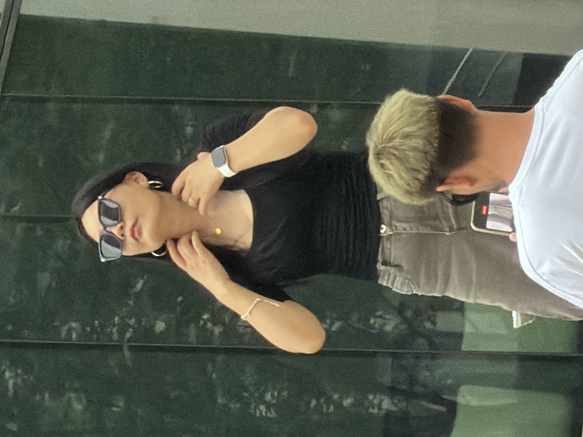 A person with dark hair, sunglasses, and hoop earrings stands against a green, paneled background. They are wearing a black top, grey pants, a white smartwatch, and a delicate bracelet. Their hands are near their neck and collarbone. In the foreground, another person, seen from the back, has dyed blonde hair and is wearing a white t-shirt. They are holding a smartphone, appearing to take a photo of the first person. The scene is outdoors, possibly in Yuzhong District, China, with reflections visible on the green panels.