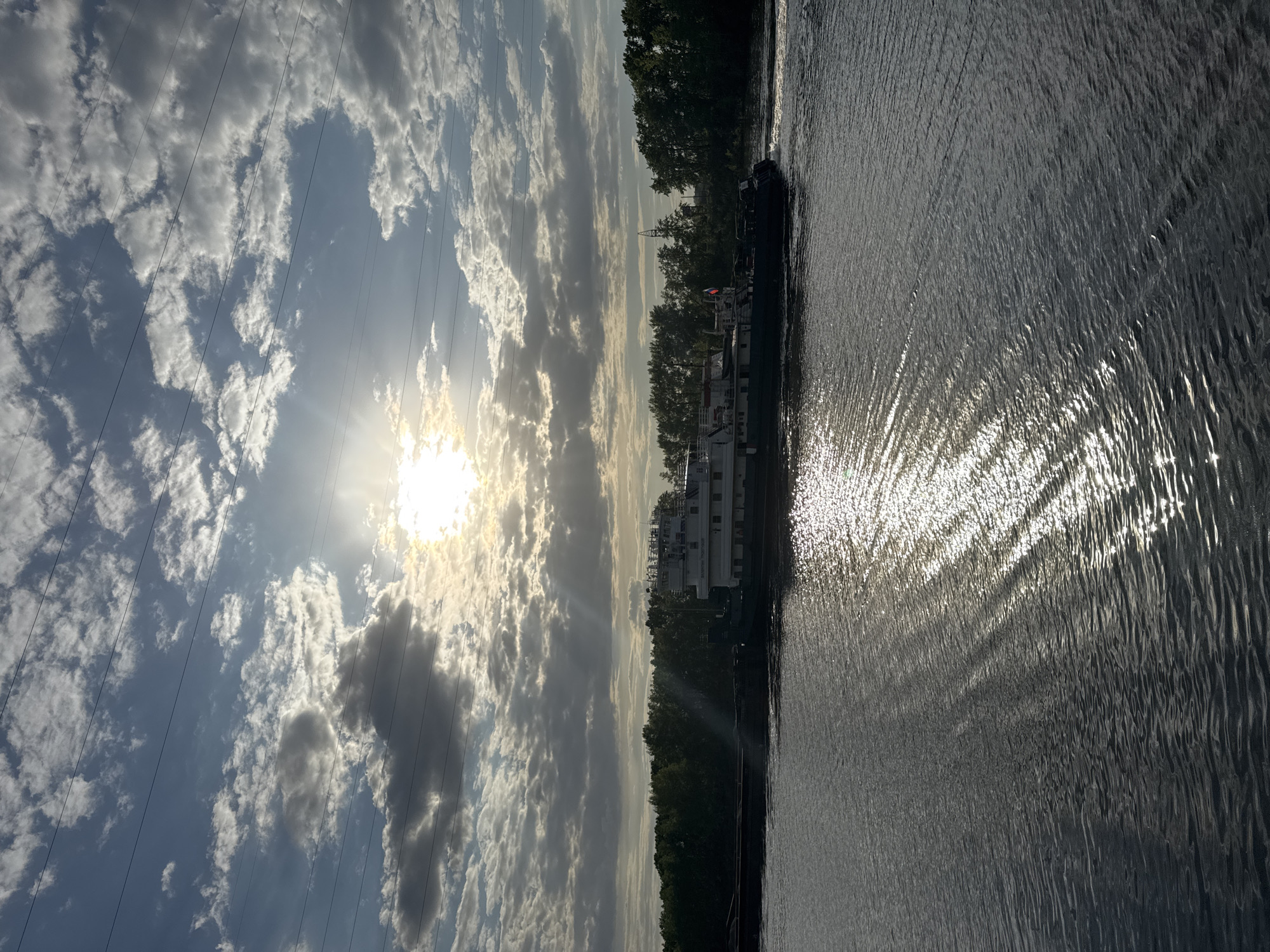 The image shows a body of water, likely a river or reservoir, reflecting sunlight. Along the riverbank, a barge-like vessel is docked, adjacent to a line of trees. Overhead are clouds in the sky, with the sun partially obscured. Electrical power lines are visible stretching across the sky. The location appears to be near the city of Chaikovsky, Russia, based on the presence of the barge-like vessel often seen on Russian waterways.