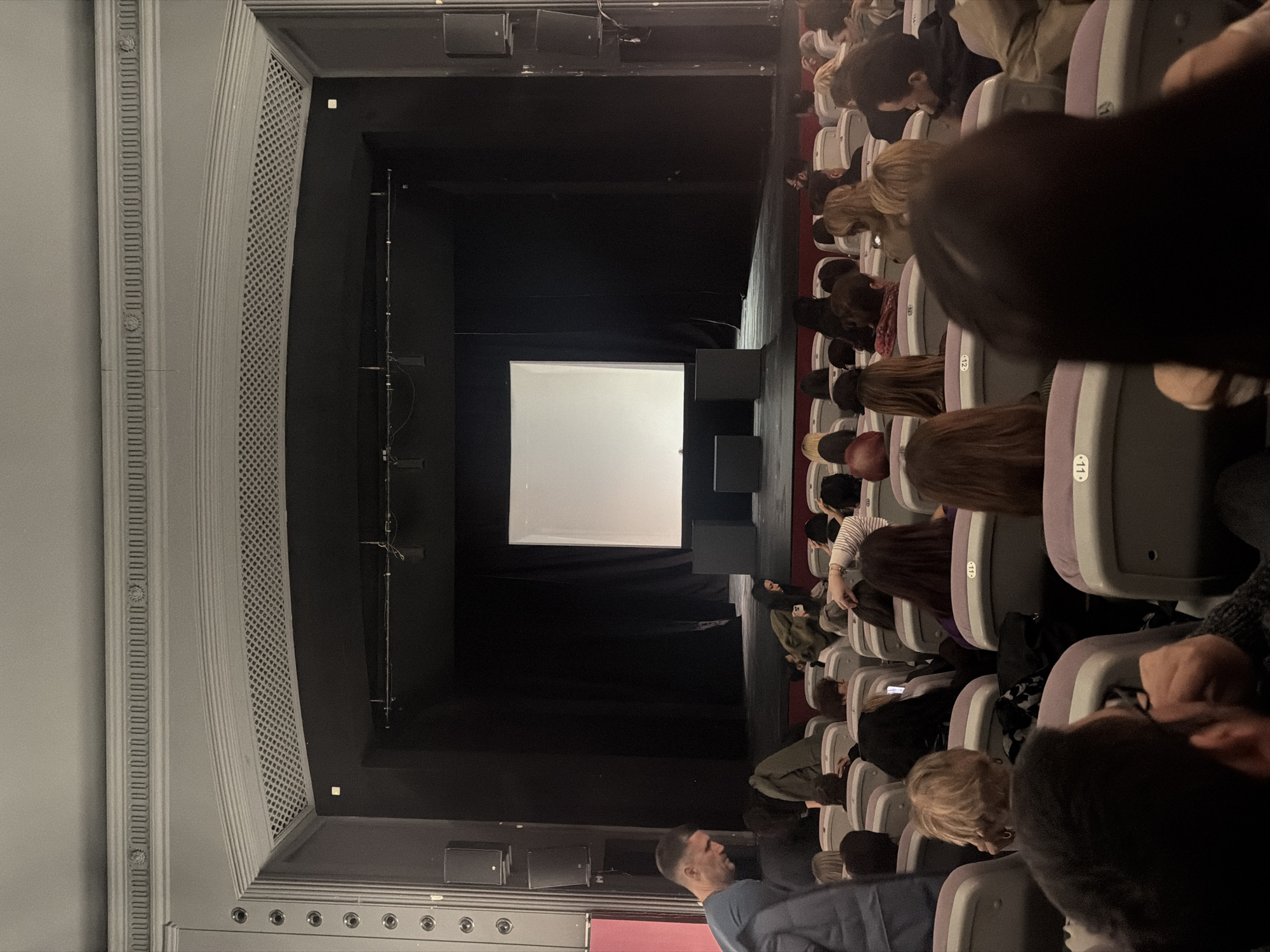 The image depicts the interior of a movie theater in Sofia, Bulgaria, filled with an audience watching a screen. The screen, located at the front, is white and illuminated. Rows of occupied seats are visible in the foreground, and audience members can be seen facing the screen. Overhead speakers are mounted on the theater ceiling.
