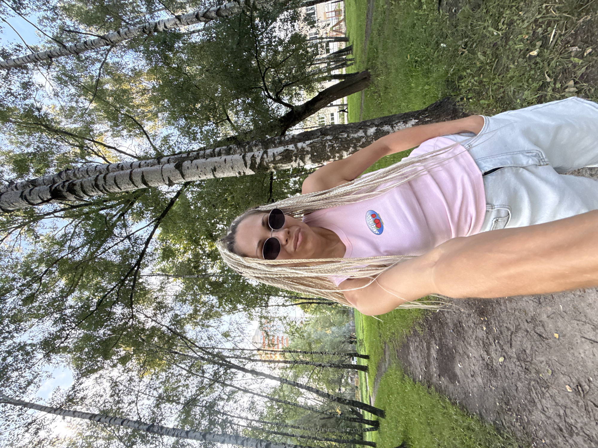 In Rusanovo, Russia, a woman with blonde braided hair and dark sunglasses takes a selfie in a park setting. She is wearing a pink tank top with a heart logo and light-colored denim shorts. The background features a dirt path, lush green grass, and several tall birch trees with white and black bark. In the distance, a multi-story building and a children's playground are visible. The sky is blue with some clouds, and the sun is shining, casting dappled light through the leaves of the trees.