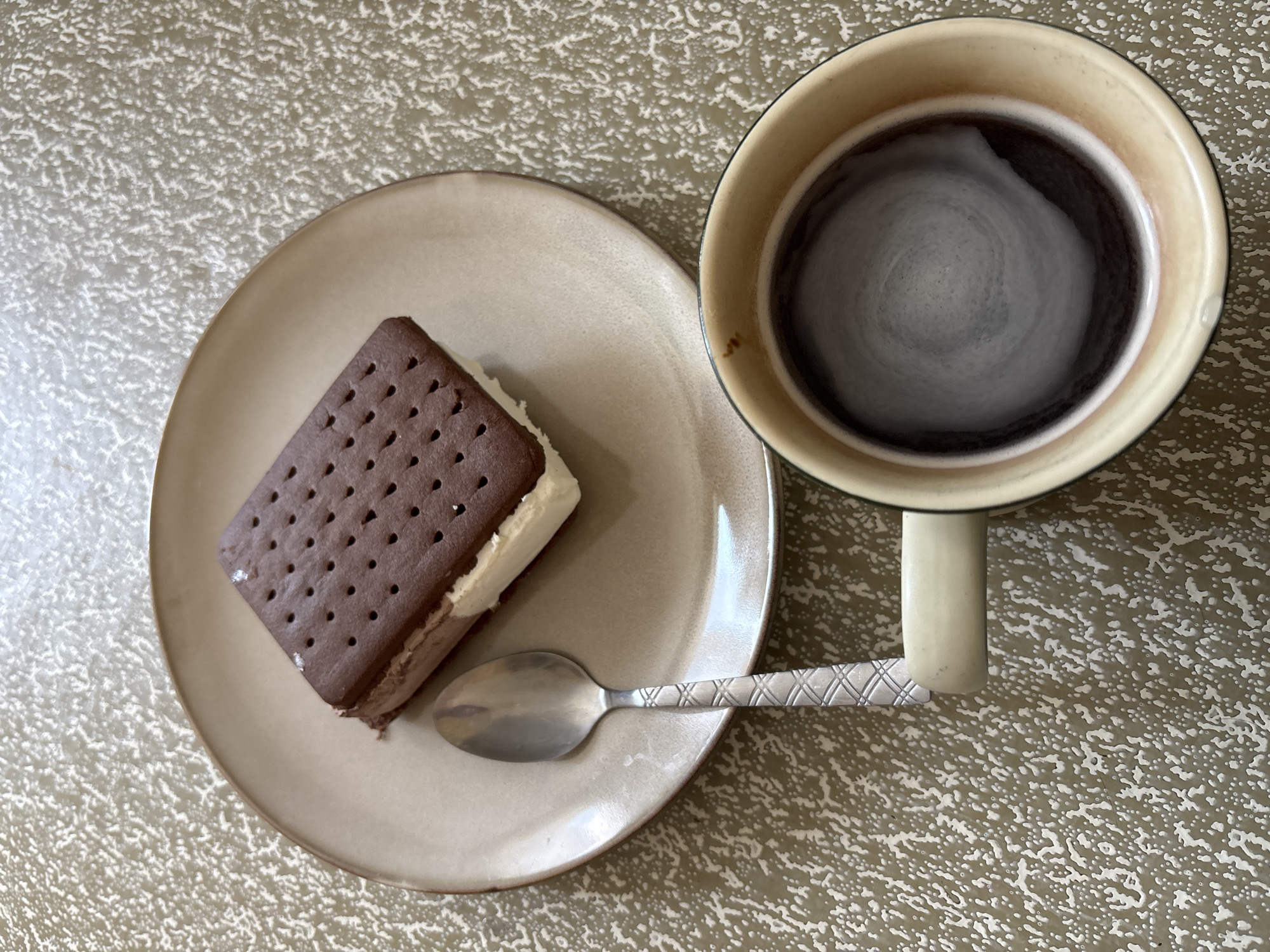 An ice cream sandwich and a mug of coffee sit on a textured, speckled surface. The ice cream sandwich is rectangular, with dark chocolate wafers and a white ice cream filling. It rests on a light brown, circular plate. A silver spoon with a patterned handle lies on the plate to the left of the sandwich. The mug is a pale yellow color with a brown rim and contains dark coffee with a foamy swirl on top. The scene is shot from a high angle, looking directly down. The environment appears to be indoors, on a table or countertop. There are no people visible in the image. The location is not specified within the image itself.