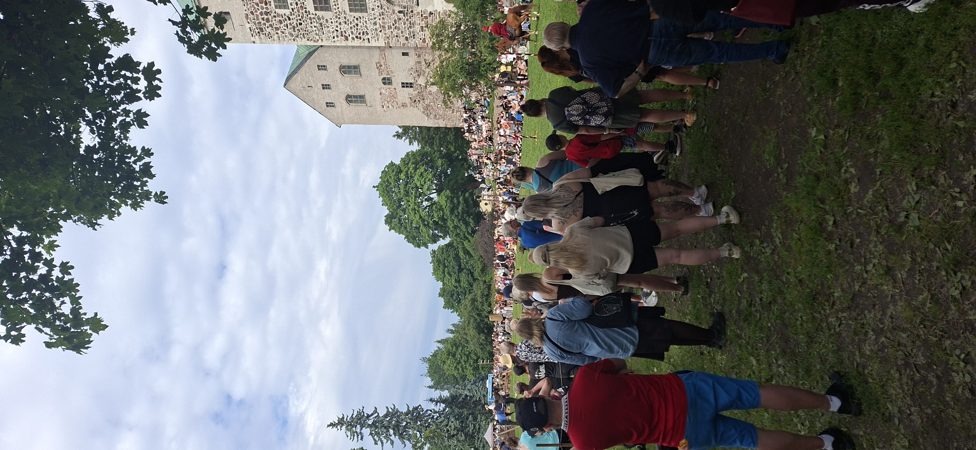 This image depicts a crowded outdoor event on the grounds of Turku Castle, Finland. A large gathering of spectators are seated and standing on a grassy field, facing towards a central performance area. In the distance, a person on horseback, wearing a red garment, is visible, suggesting a historical reenactment or performance. The castle itself, a prominent stone structure with a green roof, forms a backdrop to the event. Various trees and foliage surround the area, indicating a park-like setting within the city. The sky is overcast with scattered clouds. The foreground shows a section of the crowd, with individuals dressed in casual summer attire.