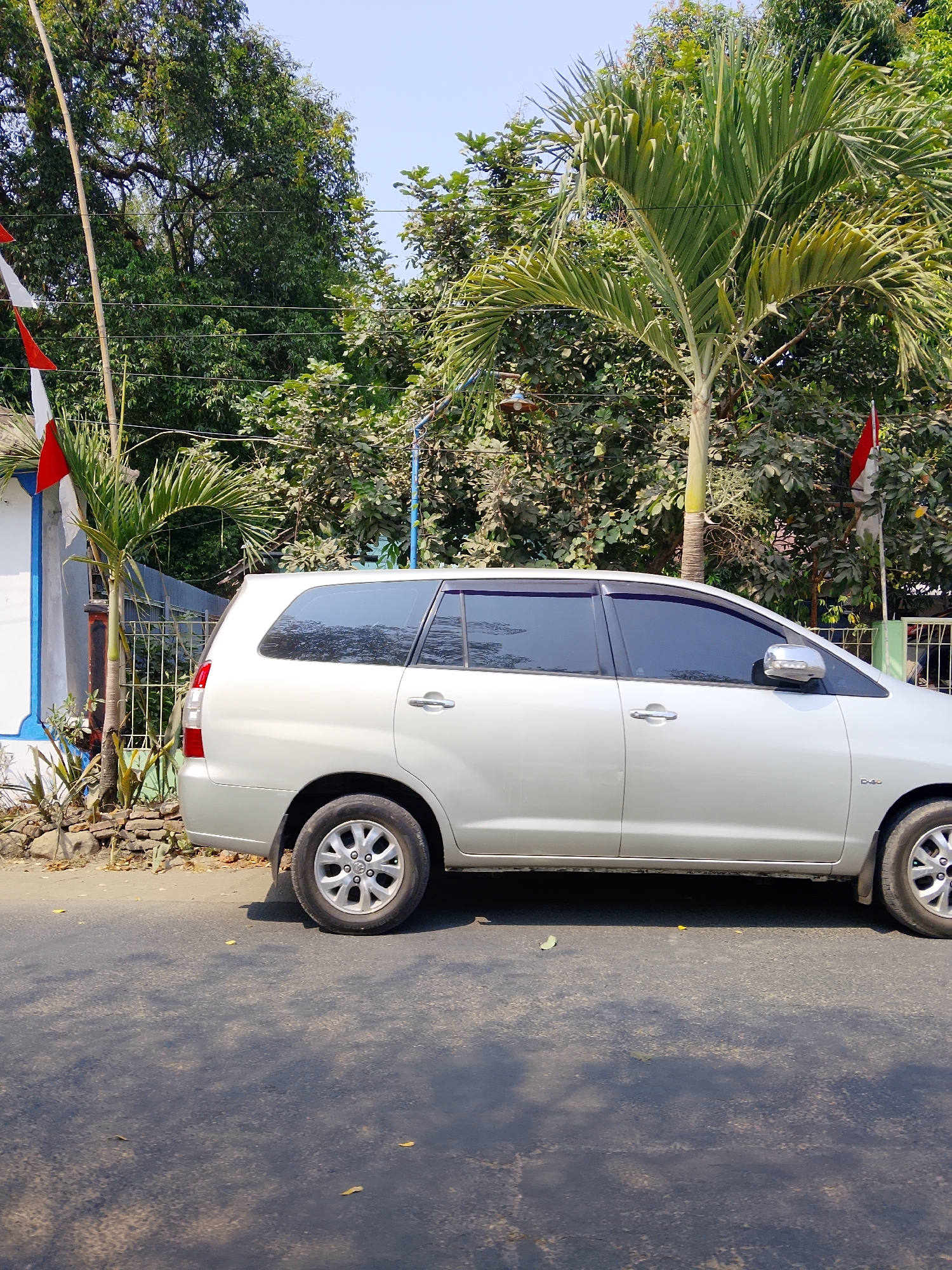 A silver station wagon is parked on the side of a paved road. The car is facing right and its side is visible. The car is surrounded by lush green trees and vegetation. There are two flags, red and white, visible in the background, possibly indicating a location in Indonesia. The sky is clear and blue. The scene appears to be a sunny day. The image was taken in the city of Ngelo, Indonesia.