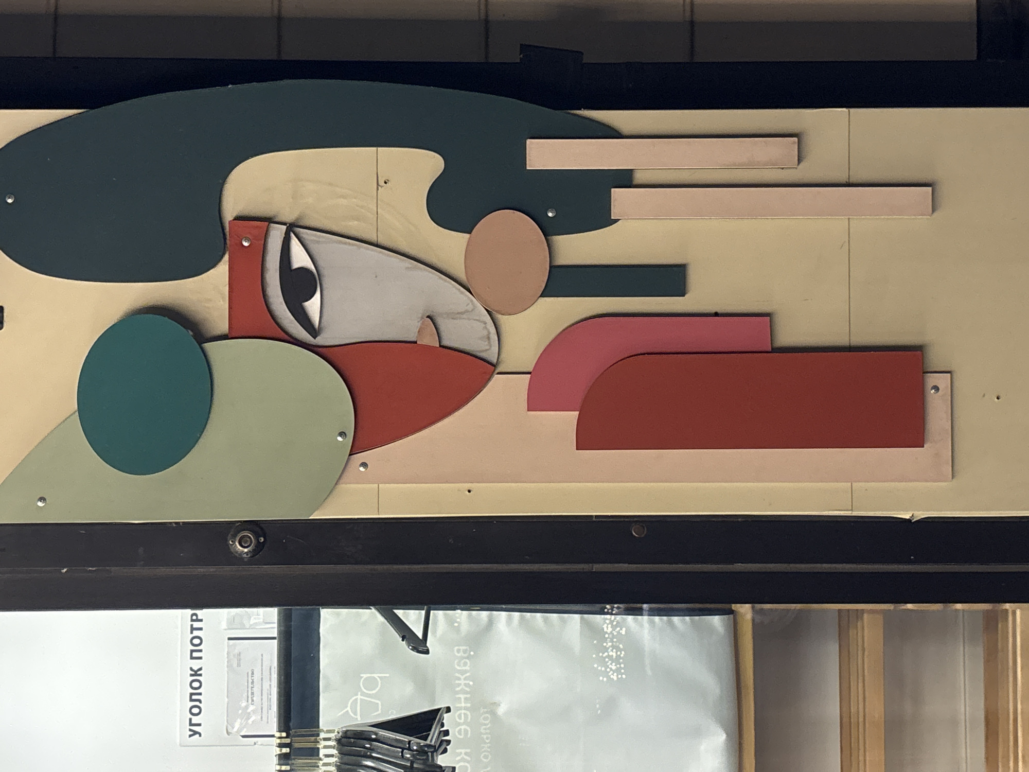 The image shows a close-up of a colorful abstract art installation featuring geometric shapes and a stylized human face. The art is mounted on a light beige background, possibly a wall or a display panel, with some vertical dark brown elements on the left and right edges, suggesting it might be a doorway or part of a facade. The artwork incorporates shades of green, red, pink, and a light beige. A prominent feature is the face, with a single eye rendered in black and white, and a gray nose and lips. Various shapes, including circles, rounded rectangles, and elongated rectangles, are layered and attached to the background, creating a sense of depth.
On the left side of the image, a dark vertical element frames a glimpse into a store. Inside, hangers with clothing are visible, along with a white bag featuring a logo that appears to be a stylized cup or container. Text in Russian is also visible on a white sign, which reads "УГОЛОК ПОТР" (Ugolok Potr), likely part of a store name or section title like "Consumer Corner." Below this, another document with Russian text, possibly a certificate or warranty, is partially visible. The floor inside the store appears to be made of light wooden planks.
The art installation and store interior suggest a retail or public space environment. Given the text in Russian and the likely retail context, the location is presumed to be a store in Novaya Usman', Russia. The overall scene is well-lit, with a slight shadow cast on the right side of the art piece, indicating a light source from the left or front.