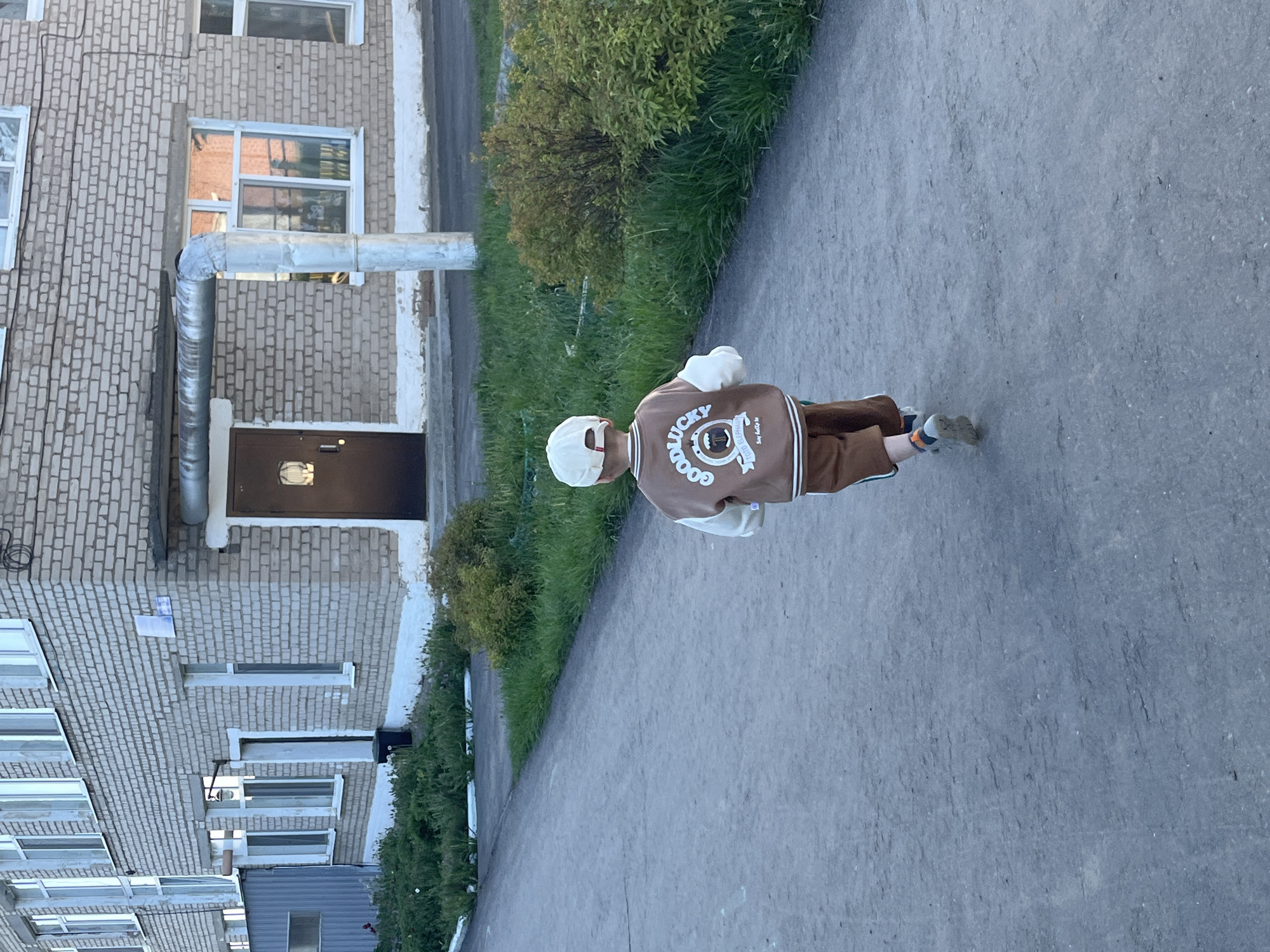 A person wearing a brown jacket and white cap is standing on a sidewalk in Фоки, Russia. The person is facing away from the camera. Behind the person is a brick building and some greenery. The sidewalk is gray. The time of day appears to be daytime.