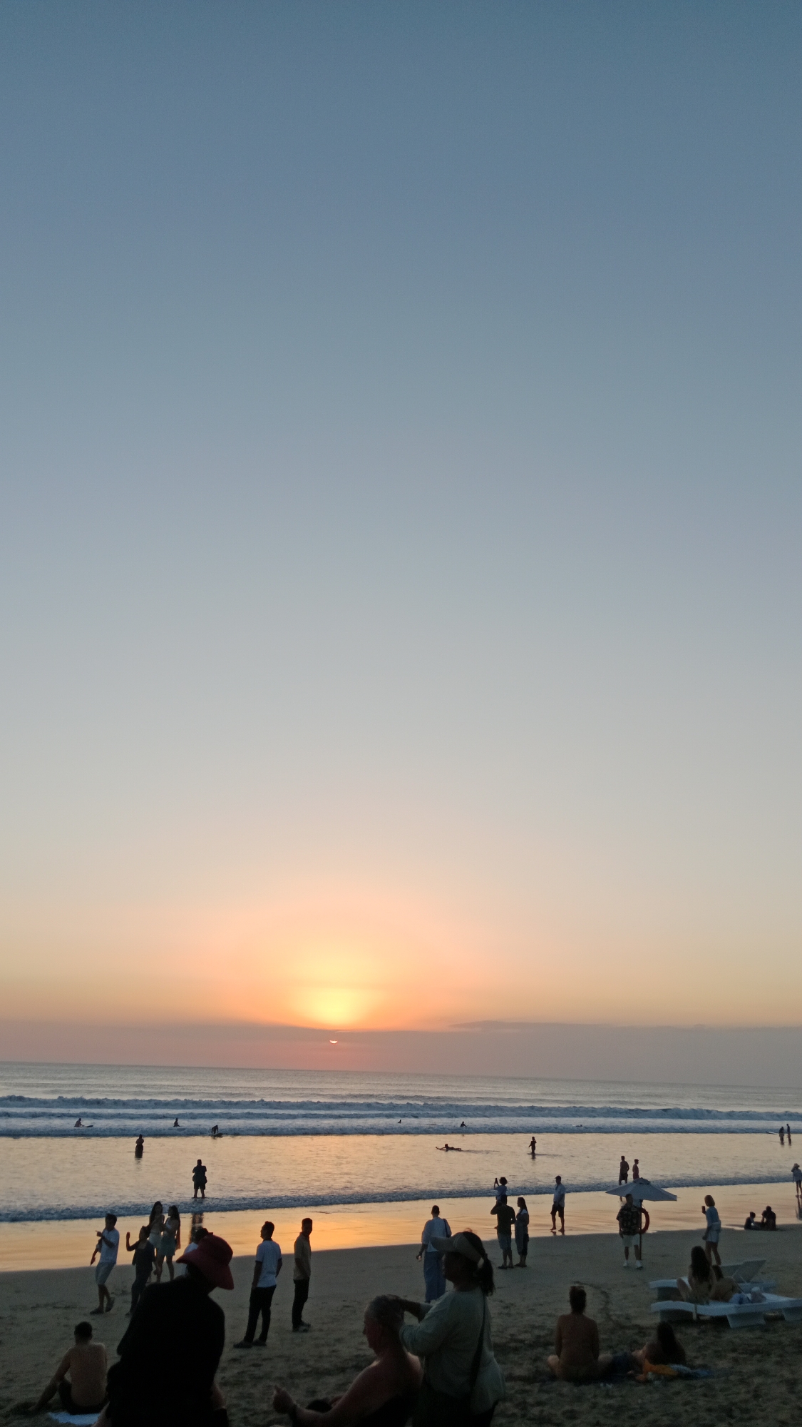 The image captures a sunset scene at Kuta Beach in Kuta, Indonesia. The sky transitions from a clear, pale blue at the top to hues of orange and pink near the horizon where the sun is setting. The sun itself is a bright orb partially obscured by the horizon line, casting a warm glow on the water. Gentle waves roll onto the sandy shore, with several people visible in the ocean, some appearing to be swimming or surfing.
On the beach, a diverse group of people are scattered across the sand. Some are standing and conversing, while others are seated or lying down, enjoying the evening. There are individuals walking along the shoreline, and some are gathered in small groups. The overall atmosphere suggests a relaxed and social environment. Beach furniture, such as lounge chairs and an umbrella, can be seen near the water's edge. The sand appears to be damp near the waterline and dry further inland. The scene is illuminated by the soft, fading light of the sunset.