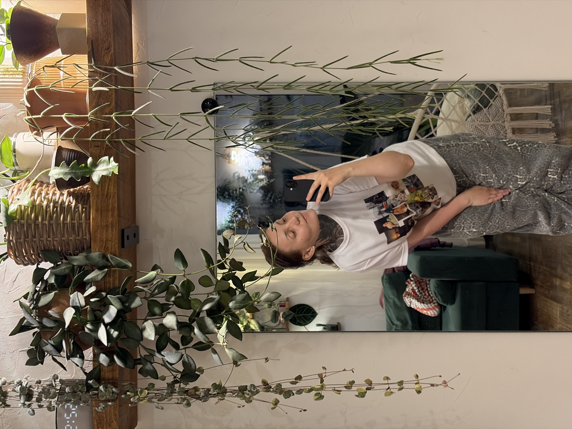 The image depicts a young woman taking a selfie in a room. She is wearing a white t-shirt with a collage of photographs printed on it and patterned grey pants. The woman has visible tattoos on her neck. She is holding a black smartphone in her right hand to take the photo, and her left hand is resting on her hip.
The background of the image reveals a mirror reflecting more of the room. In the mirror, a dark green couch with a patterned red and white throw blanket is visible. A macrame hanging chair with cushions is also seen in the reflection. The room appears to be decorated with several hanging plants, some with long, trailing vines and others with fuller foliage. A wooden shelf above the mirror holds various potted plants and decorative items, including a wicker basket. The overall lighting suggests an indoor setting, possibly a home or studio. The location is identified as Malyi Bukor, Russia.