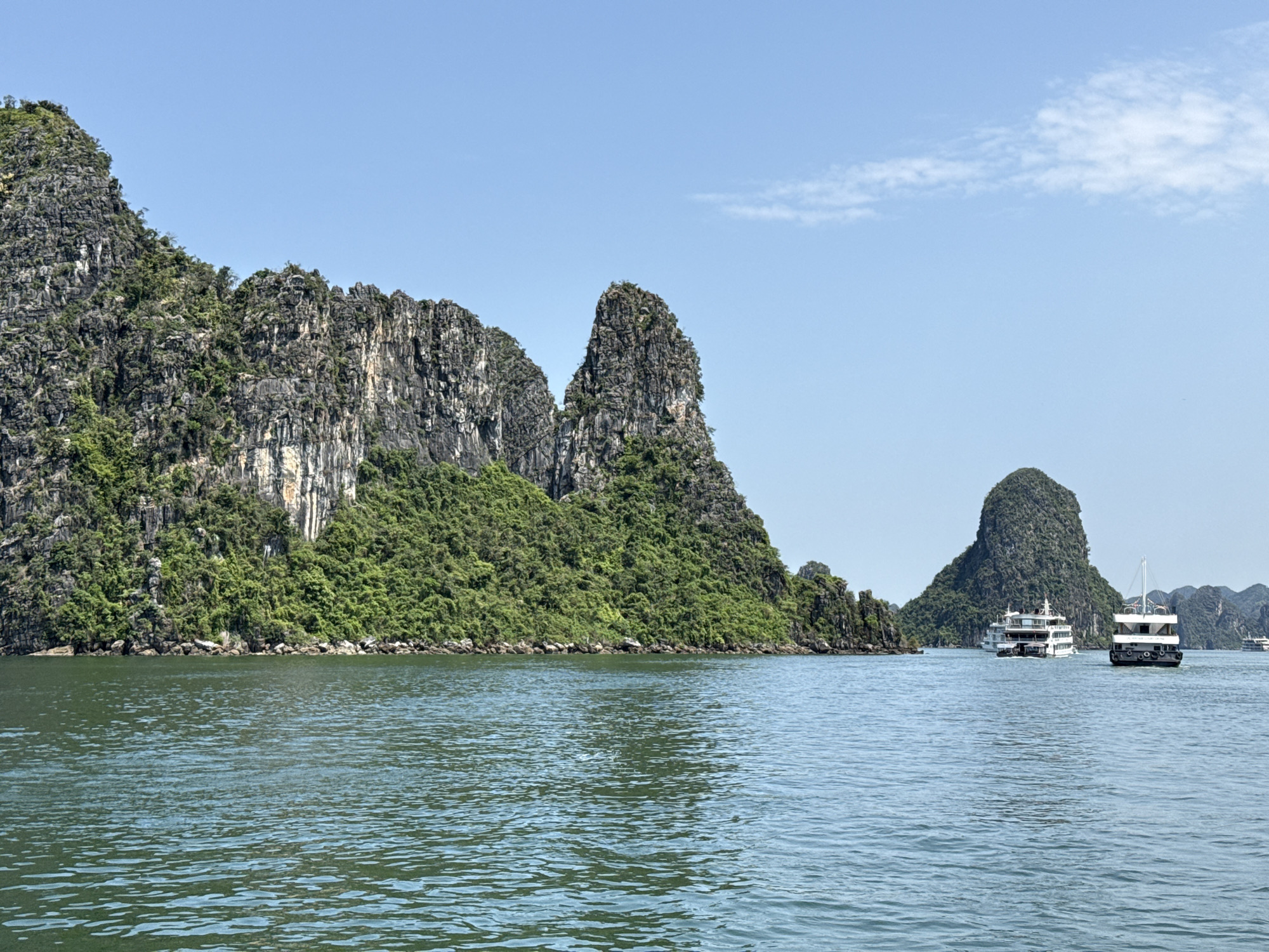 This image captures a scenic daytime view of Hạ Long Bay, near Tuan Chau Ward, Vietnam. The foreground features calm, greenish-blue water, hinting at gentle ripples. On the left and extending across the mid-ground, majestic limestone karsts rise dramatically from the bay. These towering islands are heavily adorned with dense green vegetation, contrasting with the grey, rugged rock faces.
The clear, bright blue sky with a few wispy white clouds suggests a sunny day, likely late morning or early afternoon. In the mid-ground on the right, two multi-deck boats are visible. The closer boat has a white superstructure and a dark hull, seen from its stern. Behind it, a larger, predominantly white cruise ship with dark windows is navigating through the water, with another verdant karst island in the background. The presence of these vessels indicates active tourism or sightseeing in the bay. No visible text is present in the scene. The overall impression is one of natural beauty and tranquility.