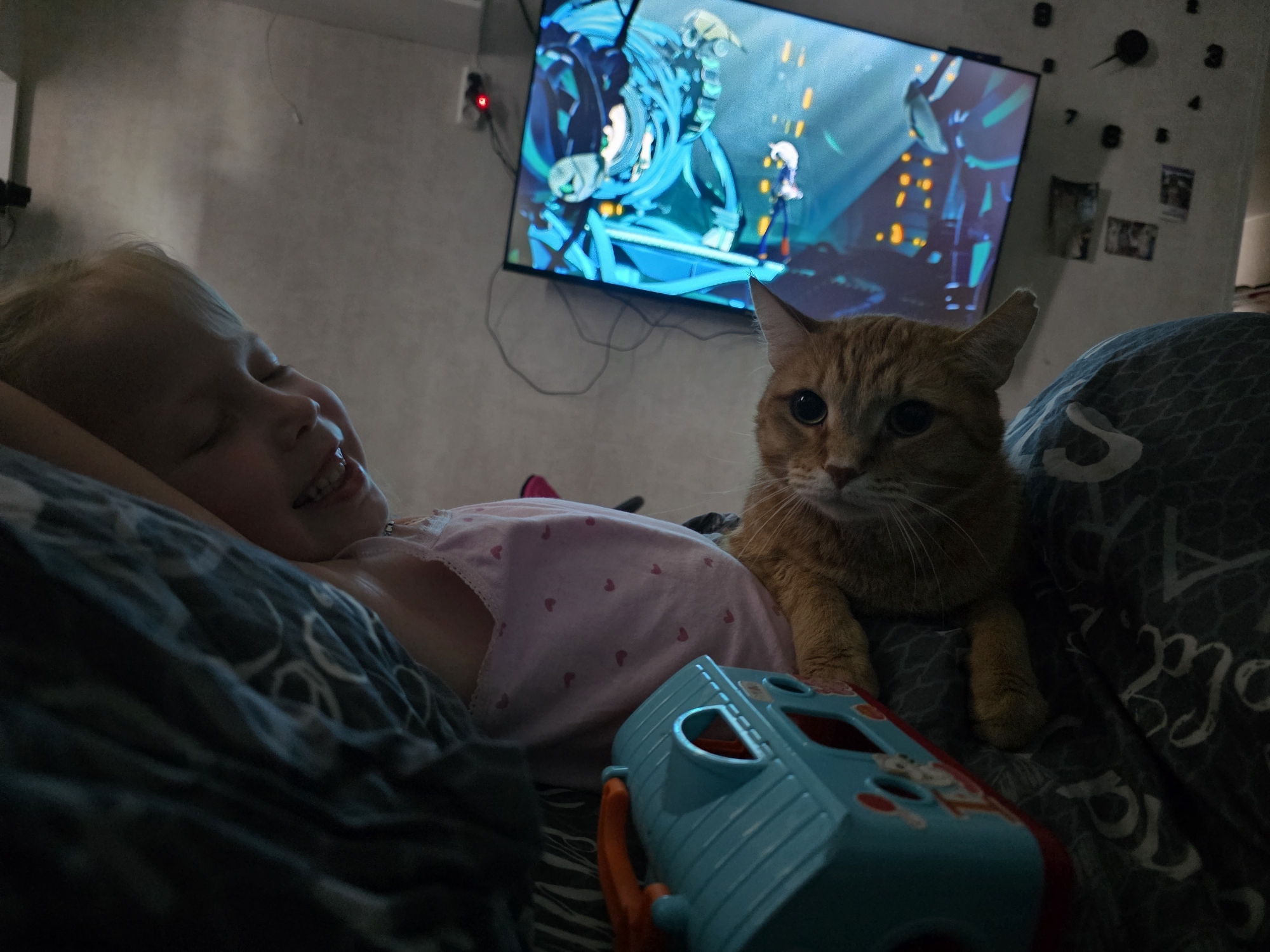 An indoor scene within a residential setting in the city of Chaikovsky, Russia, depicts a child and an orange tabby cat in close proximity, with a television visible in the background.
A young, light-haired child lies on their left side, positioned horizontally in the left-center of the frame, partially covered by dark patterned bedding. The child wears a light pink garment adorned with small red heart patterns. The child's face is turned towards the right, exhibiting a smile with eyes partially closed.
To the right of the child, an orange tabby cat sits or lies on the bedding, facing directly towards the viewer. Its pupils are dilated, and its front paws are visible.
In the background, a flat-screen television is mounted on a light-colored wall, displaying an illuminated animated scene with prominent blue and black figures and yellow architectural details. Wires are visible descending from behind the television. To the left of the television, an electrical outlet with a red indicator light is visible. To the right of the television, a wall clock composed of individual numbers (7, 8, 9, 10, 11, 12) adhered to the wall surrounds a central mechanism. Below the clock, several small, rectangular photographs or framed items are affixed to the wall.
In the foreground, a light blue and orange plastic toy, possibly a shape sorter, rests on dark patterned bedding. The overall scene is dimly lit, with the television screen serving as a significant light source.