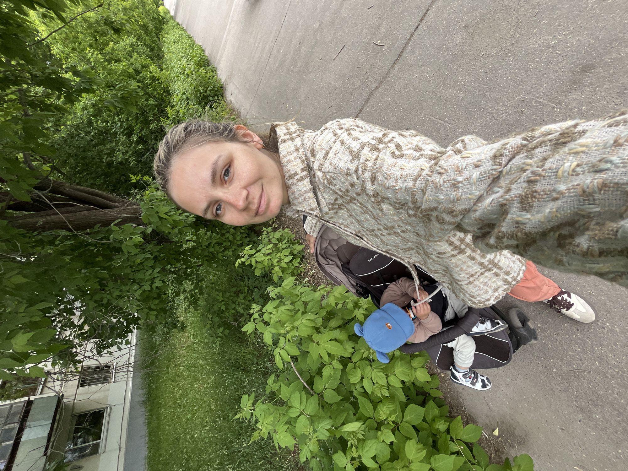 A woman stands on a sidewalk in what appears to be Chaikovsky, Russia, judging by the visible building and vegetation. She is taking a selfie while holding a baby stroller. The stroller contains a young child wearing a blue hat and light-colored clothing, playing with a white object. The woman is wearing a woven jacket and pants, and contrasting sneakers. The environment includes green bushes and trees along the sidewalk, as well as a building facade visible in the background. The sidewalk is paved.
