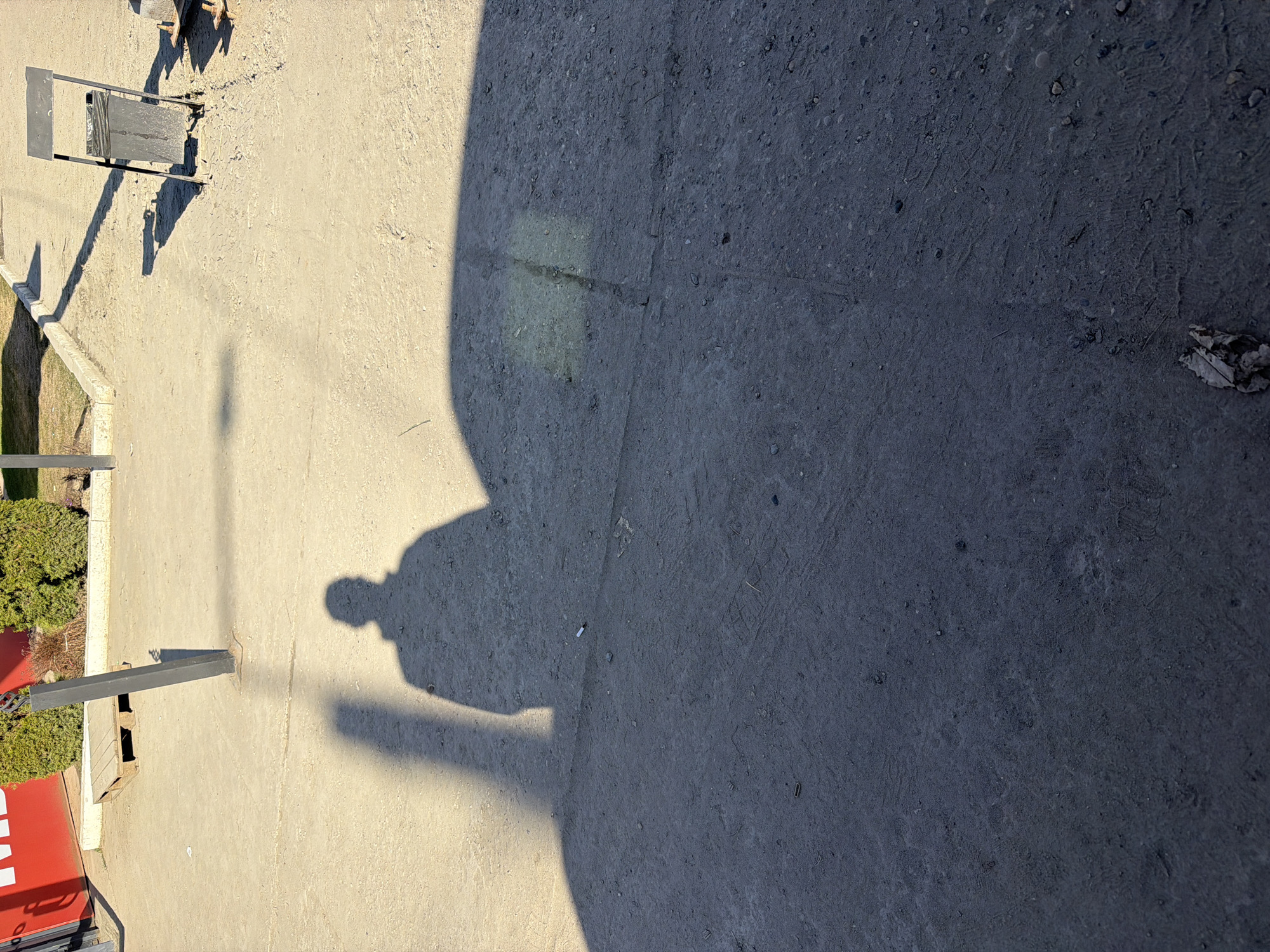 An overhead, high-angle shot captures an outdoor scene dominated by strong shadows on paved ground. The image is oriented vertically, with subjects appearing sideways.
The ground is split into two distinct areas: a lighter, sandy-colored concrete or asphalt area on the left and a darker, coarser-textured gray asphalt or dirt area on the right. A large, dark, amorphous shadow, possibly from a building or a large overhead structure, covers much of the darker ground on the right.
Prominently, a human shadow, clearly defined with a head, torso, and legs, is cast across the lighter pavement, extending towards the darker ground. Its long, distinct form suggests the sun is low in the sky, indicating either early morning or late afternoon. Another long, narrow shadow emanates from a black lamppost positioned on the lighter ground. Nearby, a metal frame structure, possibly a stand for a trash bin or a sign, is also present, casting its own shadow. Small green bushes are visible in the background along the edge of the paved area on the left.
Notable details include small pebbles and dirt on the ground, a discarded cigarette butt near the human shadow, and a dried leaf on the darker pavement. On the very bottom left, a fragment of a red sign with white lettering is visible, showing parts of letters that appear to be "MA". The overall impression is a sunny, clear day in an urban or suburban outdoor setting.
