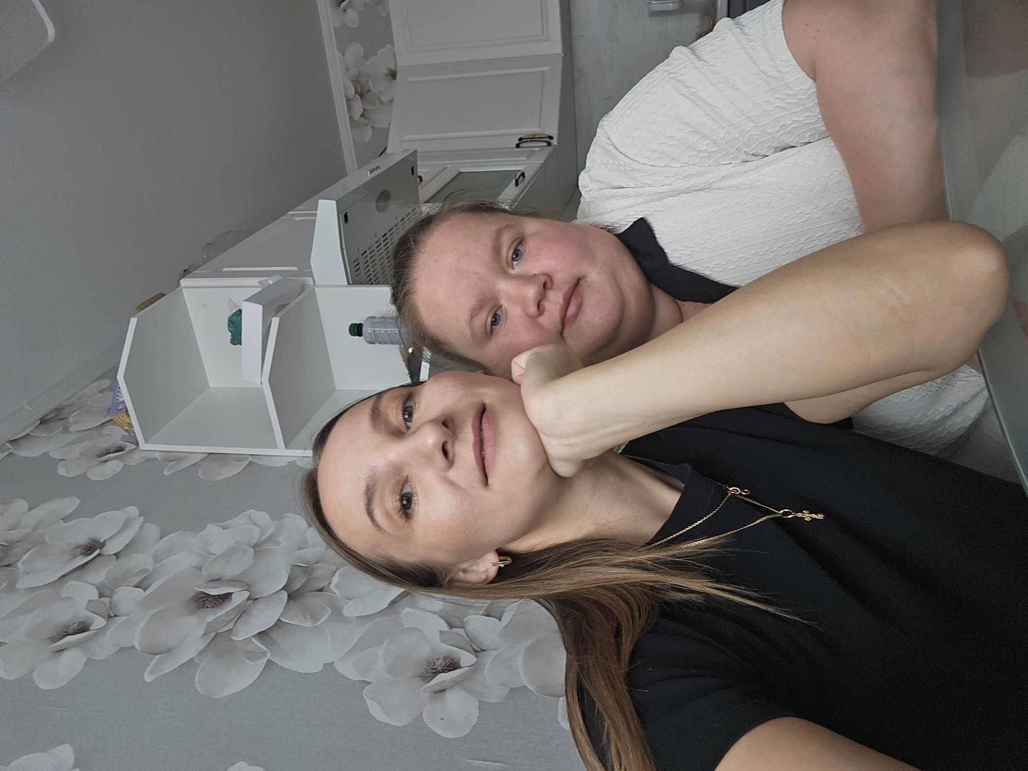 Two women are positioned close together, likely indoors based on the wallpaper with floral patterns. The woman in the foreground has brown hair, is wearing a black top, and has a gold necklace. Her hand is placed on the cheek of the other woman. The woman in the background has blonde hair and is wearing a white top. The scene includes a white shelf unit and kitchen cabinets in the background. Without additional information or context, it is impossible to definitively confirm that this picture was taken in the city of Chaikovsky, Russia.