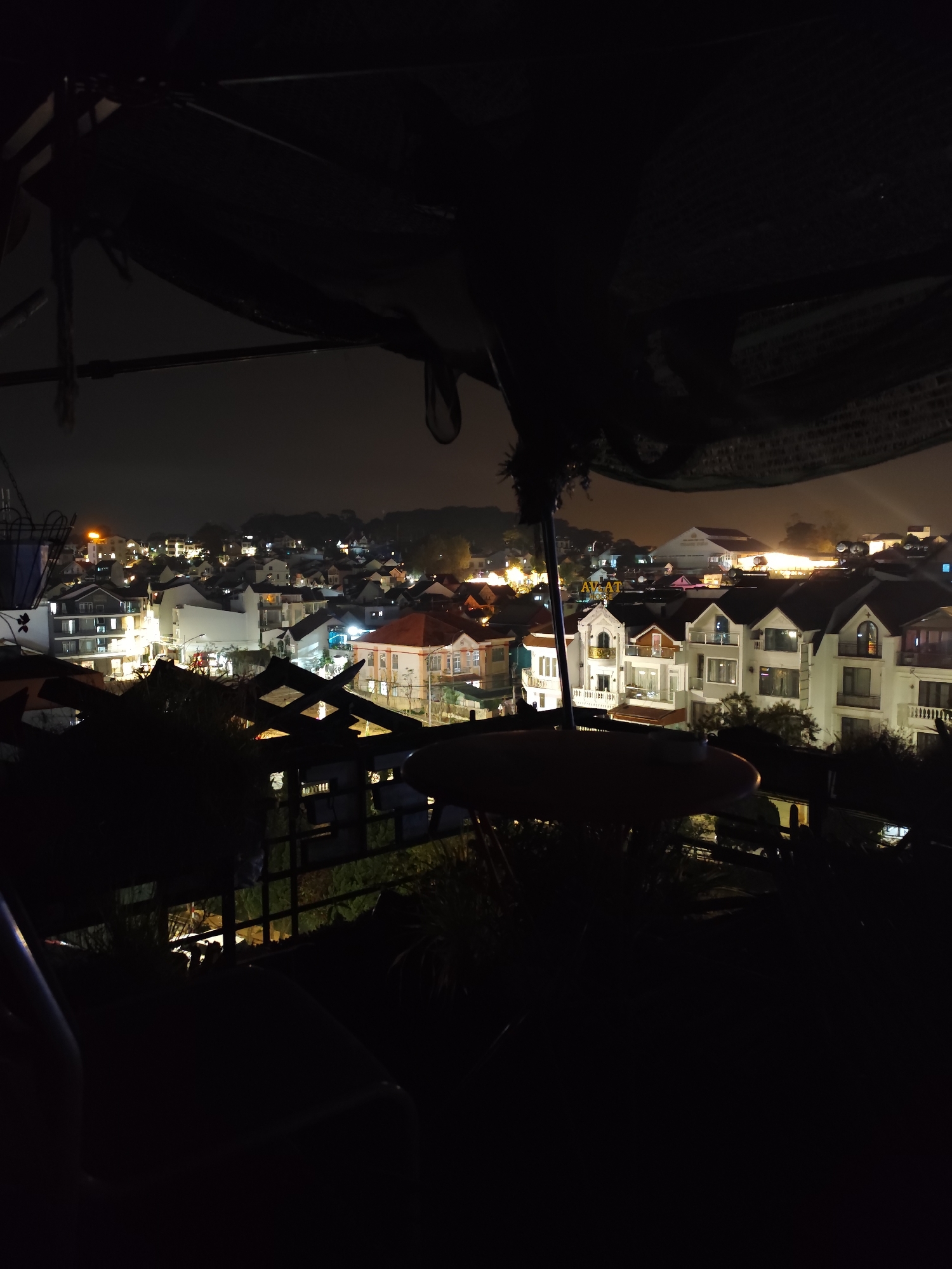 A nighttime, elevated view shows buildings in Da Lat, Vietnam. The foreground is dark with a partial view of a canopy or shade structure. In the midground, numerous illuminated buildings are visible, displaying a variety of architectural styles, some with red-tiled roofs and white facades. Streetlights and building lights create a scattered pattern of illumination across the cityscape. The background shows a hazy, dark sky. No people are discernible in the image.