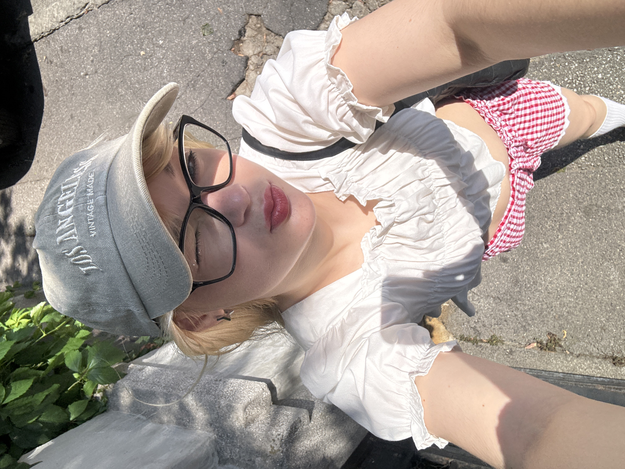 A person with blonde hair and glasses is lying on the ground outdoors. They are wearing a white ruffled top, a red and white checkered skirt, white socks, and a light grey baseball cap. The cap has text embroidered on it, but it is partially obscured. The person is looking up and their face is illuminated by sunlight, suggesting it is daytime. The ground is paved with asphalt and there are some green plants visible. The overall scene suggests a casual outdoor setting, possibly in a park or on a street. There are no other people or significant objects in the frame. The lighting and shadows indicate a sunny day.
