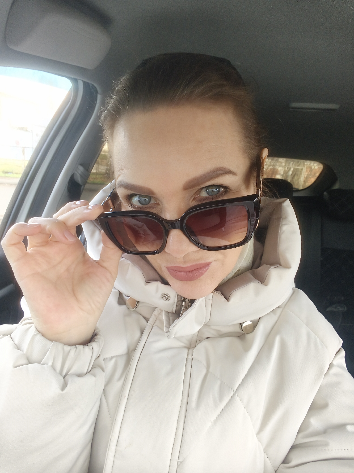 A woman is seated inside a car, looking directly at the viewer. She is holding a pair of dark, rectangular sunglasses slightly down, peeking over the top of them with light-colored (blue/grey) eyes. Her hair is pulled back, and she is wearing light-colored lipstick. She is dressed in a light-colored, possibly cream or beige, quilted puffer jacket with a high collar, which appears to be fastened with a zipper and possibly snaps.
The setting is the interior of a car, with the roof liner, sun visor, and door frame visible. Through the car windows on either side, an outdoor environment is blurred but appears to be daytime, possibly overcast, with some light foliage or buildings in the background. The lighting suggests it is daytime. Her expression is engaging and direct, conveying a sense of playful intrigue or confidence. No visible text is present in the image.