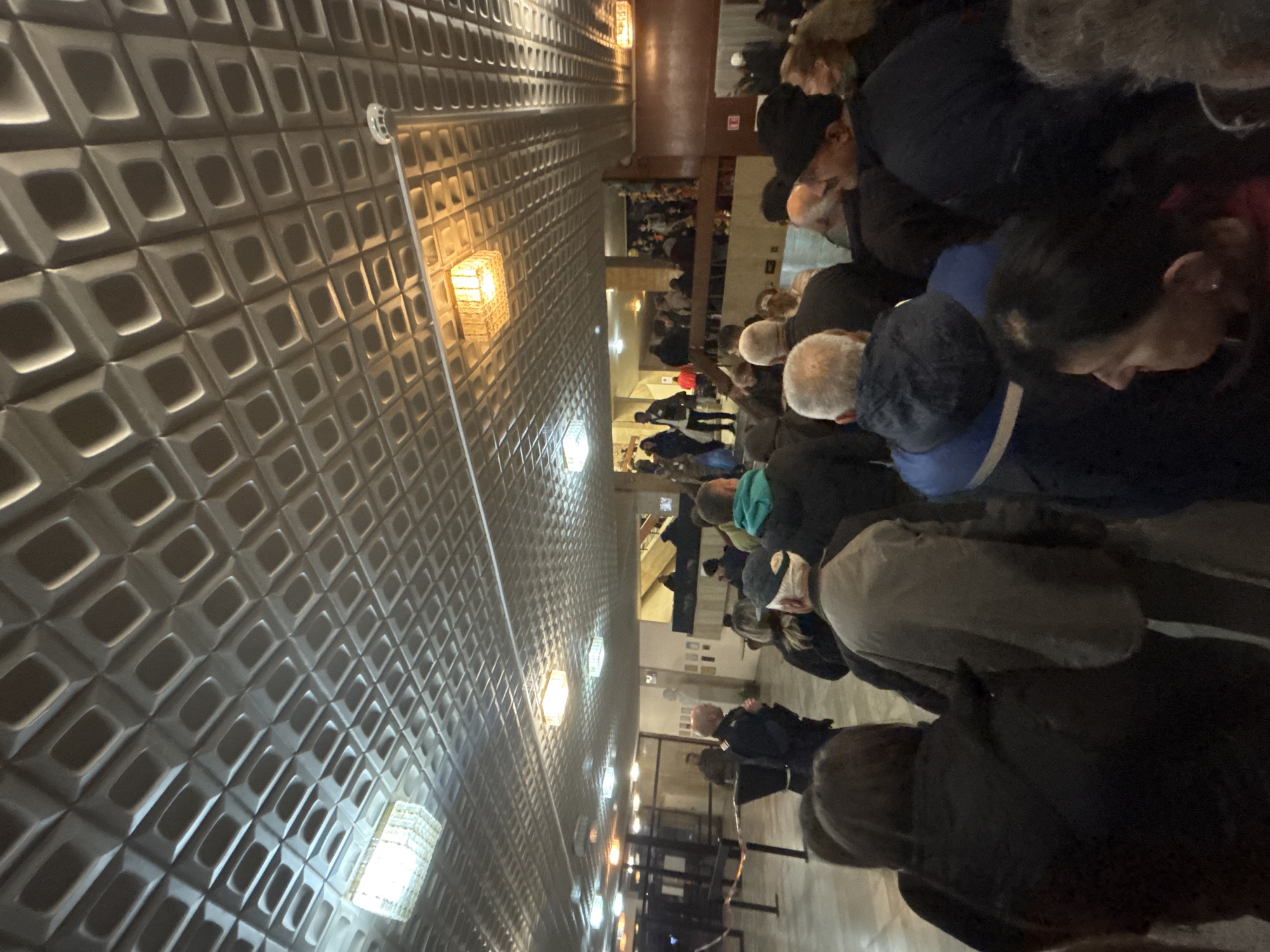The image shows a crowded indoor scene, possibly a waiting area or hallway. The walls are covered in a patterned, textured surface. Overhead lighting is present. Multiple people are visible, seemingly queuing or waiting. The location is not discernible from the image. The city of Kardzhali, Bulgaria cannot be confirmed or denied as the location.