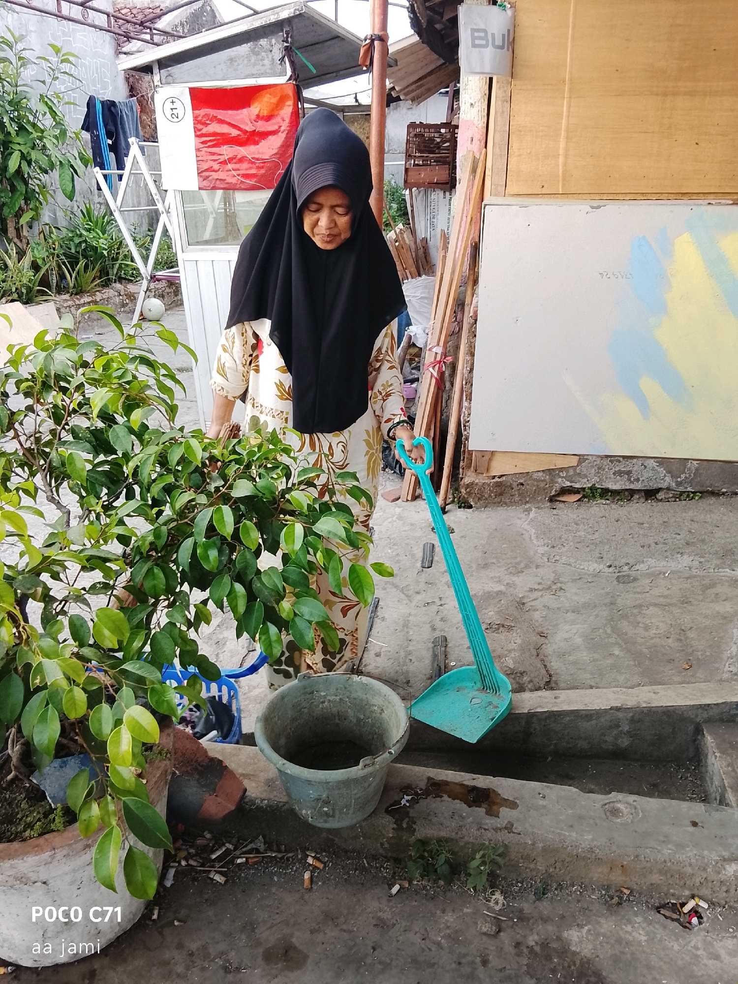 An elderly woman wearing a black hijab and a floral patterned dress is standing outdoors in what appears to be a residential area in Tasikmalaya, Indonesia. She is looking down, and her hands are near a bright turquoise dustpan and broom set. Next to her is a large green plant in a pot and a grey bucket. Behind her, a clothesline with laundry is visible, along with some building structures. The ground is concrete and shows signs of debris, including cigarette butts and discarded items. The overall scene depicts a moment of everyday life, possibly during a cleaning activity, in a semi-urban environment.