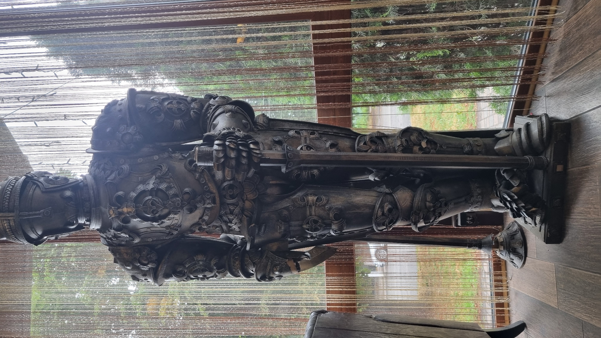 This image shows a life-sized statue of a knight in full armor, standing indoors. The statue is dark metallic, with intricate details on the armor, including decorative elements and what appear to be fleur-de-lis symbols. The knight is holding a sword vertically, its tip resting on the base. The statue is positioned in front of a large window that looks out onto an outdoor scene. Through the window, there are trees and a road with a white car visible. The window is partially covered by a beaded curtain, which hangs in vertical strands. The floor in the foreground is made of wooden planks. The scene appears to be a public or commercial space, possibly a cafe, restaurant, or shop, given the interior decor and the view outside. There is no indication of a specific location within the city of Smila, Ukraine, other than the general description of the setting.