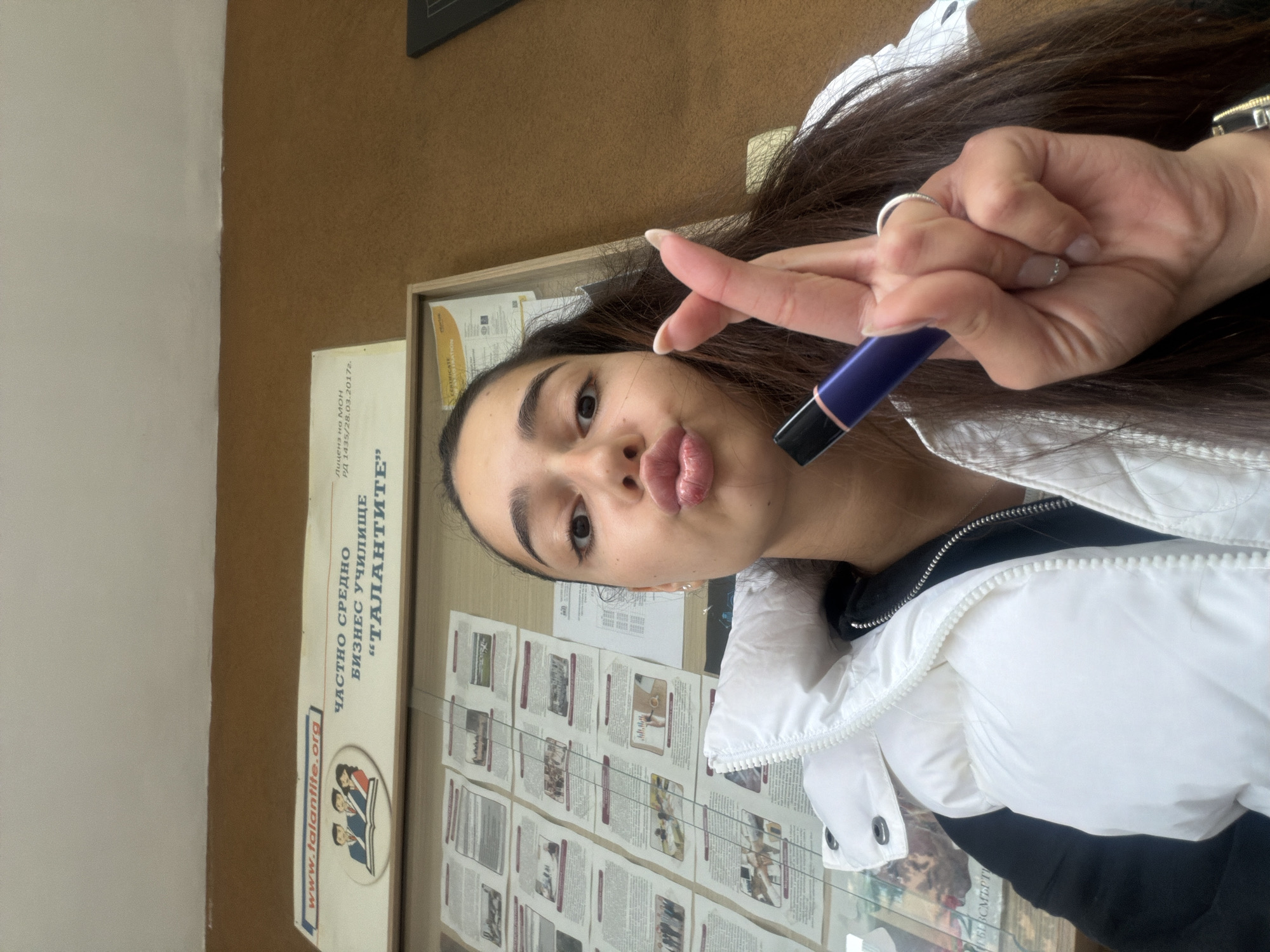 A young woman is taking a selfie with her right hand raised, holding a purple and black vape pen. She has long dark hair and is wearing a white puffer jacket over a black top. Her lips are pursed in a kissing expression. She is standing next to a bulletin board containing documents, including a sign that reads "ЧАСТНО СРЕДНО БИЗНЕС УЧИЛИЩЕ "ТАЛАНТИТЕ" (Private Secondary Business School "The Talents")". The text "www.talantite.org" and "Лиценз на мон РД 1435/28.03.2017г." (License of MON RD 1435/28.03.2017) are also visible on the sign. The location is likely in Plovdiv, Bulgaria.