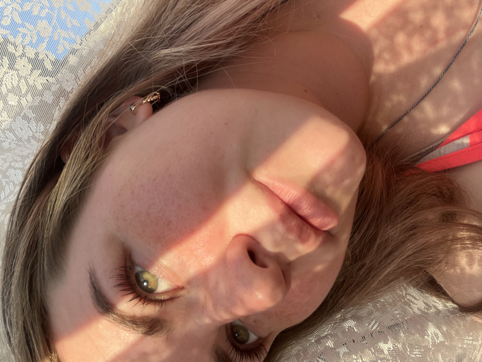 A close-up, tilted portrait shot features a young woman with light brown hair and green eyes. She lies on a patterned white surface, possibly a blanket or fabric. A beam of light crosses her face, casting shadows and highlighting her features. She wears a gold earring and a necklace with a delicate chain. Part of a pink garment is visible near her shoulder. The location is unknown and cannot be determined from the visual data, nor can the city of Chaikovsky, Russia, be confirmed.