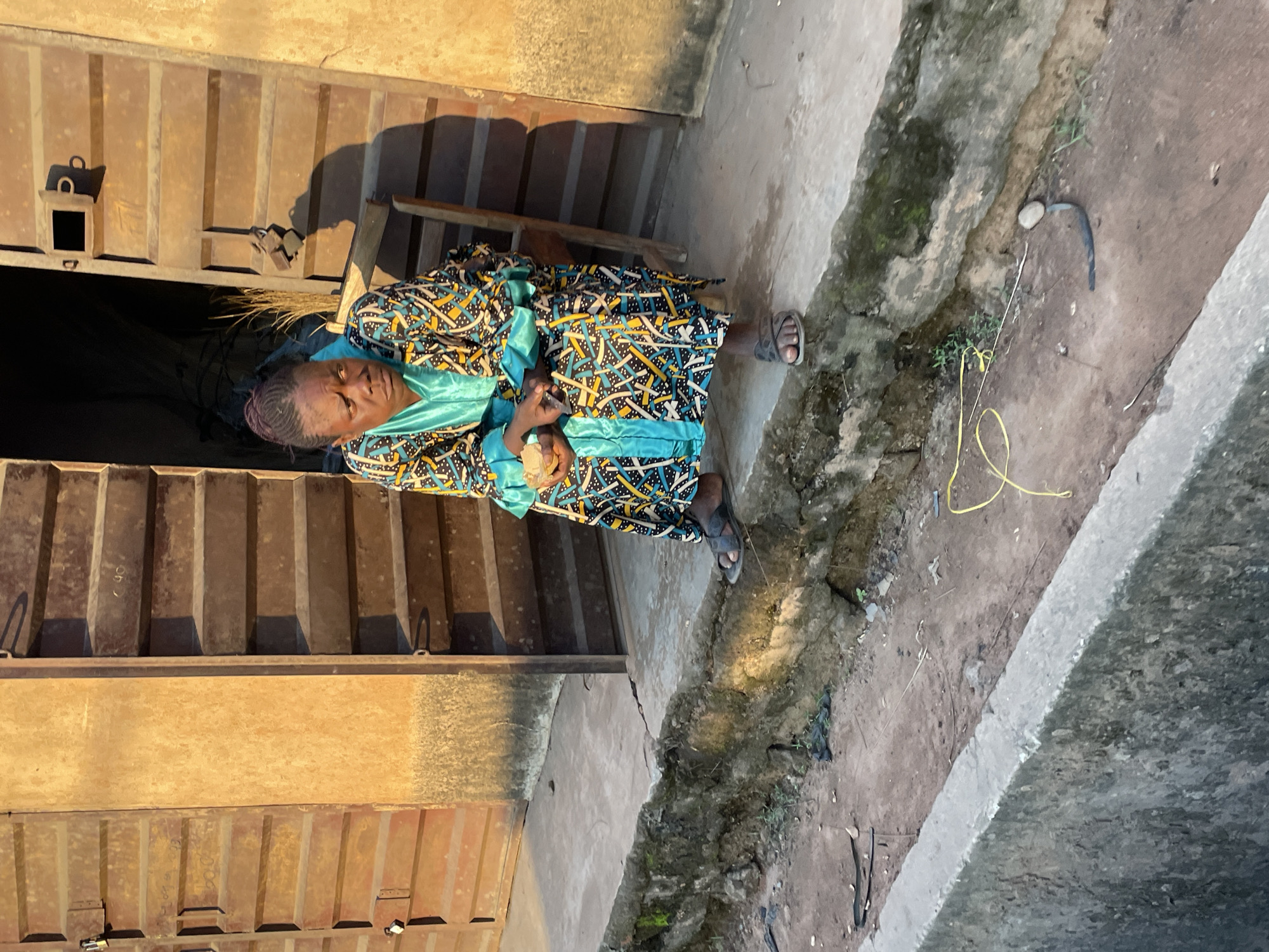 A Black woman sits on a wooden chair in front of a building with corrugated metal doors in Nkpor, Nigeria. She is wearing a colorful, patterned dress with a turquoise collar and trim, and dark sandals. Her hair is styled in braids pulled back from her face. She is looking to her left with a thoughtful expression and holding a small, light-colored object in her hands. The building's doors are weathered and show signs of age. The ground in front of the building is concrete, and to the left, there is a raised stone ledge with dirt and vegetation. A yellow cord lies on the ground near the ledge. The lighting suggests it is either early morning or late afternoon, casting shadows.