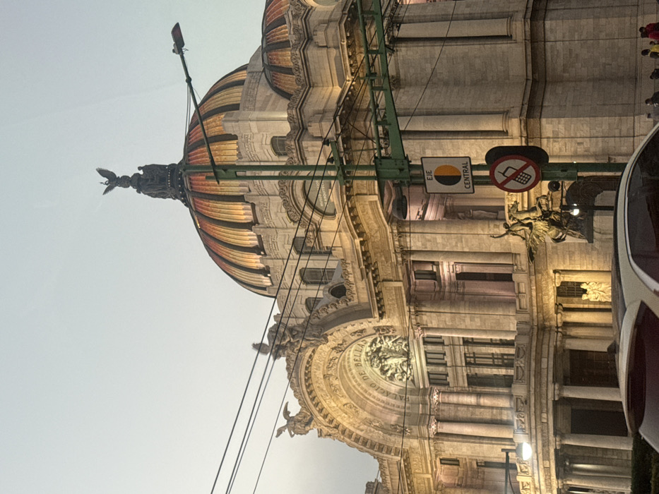 The image captures a striking view of a grand, ornate building, identified by the inscription as the "Palacio de Bellas Artes" (Palace of Fine Arts). The building is rendered in a classical architectural style, characterized by its imposing size, numerous columns, arched windows, and intricate sculptural details.
A prominent feature is the building's distinctive dome, which displays a vibrant, multi-colored pattern of striped segments in shades of gold, orange, and reddish-brown. Perched atop the dome is a large, winged statue, possibly an eagle or a similar mythological creature, adding to the building's majestic silhouette against the sky. Below the dome, the upper facade is adorned with elaborate carvings and additional statues of winged figures.
On the main facade, a semi-circular archway frames a central sculpture, and around it, the words "PALACIO DE BELLAS ARTES" are clearly inscribed, confirming the building's identity. The building's stonework appears to be a light, possibly beige or cream color, and it is illuminated by artificial lights, suggesting either dusk, dawn, or an overcast day, as the sky is a soft, light gray-blue.
In the foreground, several urban elements are visible. A green metal structure, likely part of a lamppost or traffic signal framework, spans across the image. Hanging from this structure are two street signs. One is a vertical rectangular sign with "EJE CENTRAL" (Central Axis) written on it, accompanied by a circular logo that is half yellow and half dark. The other sign is circular, featuring a red border, a white background, and a crossed-out mobile phone icon, indicating a restriction on cell phone use. Utility wires are also visible, crossing the upper portion of the frame against the sky.
On the far right side of the image, a partial view of a vehicle, possibly a tour bus or car with red and white elements, suggests the photograph was taken from a street-level perspective, perhaps from inside a moving or stationary vehicle. The overall impression is one of a significant cultural landmark in an urban setting.