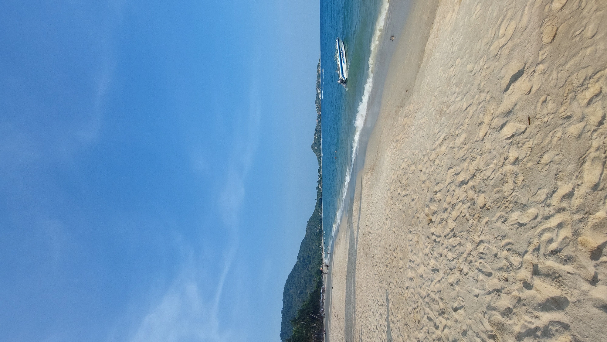 This image captures a picturesque tropical beach scene, likely in Baan Lamai, Thailand. The view is dominated by a clear, bright blue sky that occupies the upper portion of the frame, showing only a few faint, wispy clouds.
Below the expansive sky lies the calm, turquoise ocean, its surface gently rippling with small waves breaking softly onto the shore. A white speedboat with blue detailing, and what appears to be the word "PHUKET" written on its side, is partially beached or anchored very close to the waterline. In the distance, a lush, green mountainous coastline extends along the horizon, covered in dense tropical foliage. Small buildings and structures are visible nestled at the base of these mountains, indicating human presence. A few indistinct figures can be seen in the distant water or walking along the far stretch of the beach.
The lower portion of the image showcases a wide expanse of light, sandy beach. The sand is covered with numerous footprints, suggesting recent activity and movement by people enjoying the area. The bright, natural lighting indicates it is daytime, likely late morning or early afternoon, with the sun reflecting off the water and sand. The overall atmosphere is one of serenity and leisure, characteristic of a sunny day at a beautiful coastal destination.