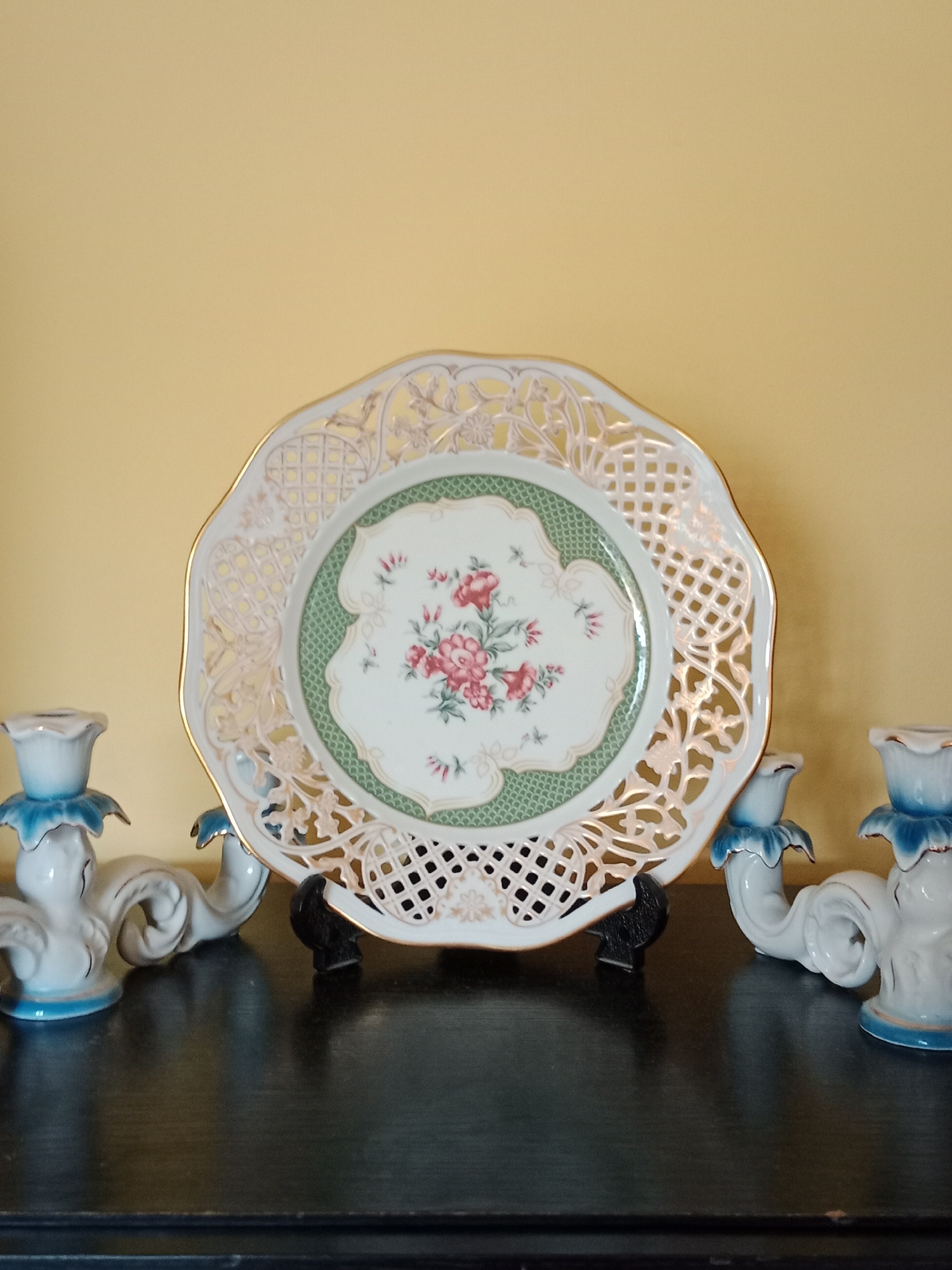 This is a still life image of decorative porcelain items arranged on a dark wooden surface against a plain yellow background. The central object is an ornate, octagonal decorative plate with a pierced lace-like border and a scalloped edge. The plate features a central floral motif with pink roses and green foliage, surrounded by a green textured border and further decorative elements. To the left and right of the plate are two matching porcelain candle holders. These candle holders are white with blue accents around the top where a candle would sit, and feature ornate, curved arms. The objects appear to be indoors, possibly in a domestic setting. There are no people visible in the image, and no actions are taking place. The lighting is even, suggesting an artificial light source. There is no information in the image that would allow for the determination of its location in Bydgoszcz, Poland.