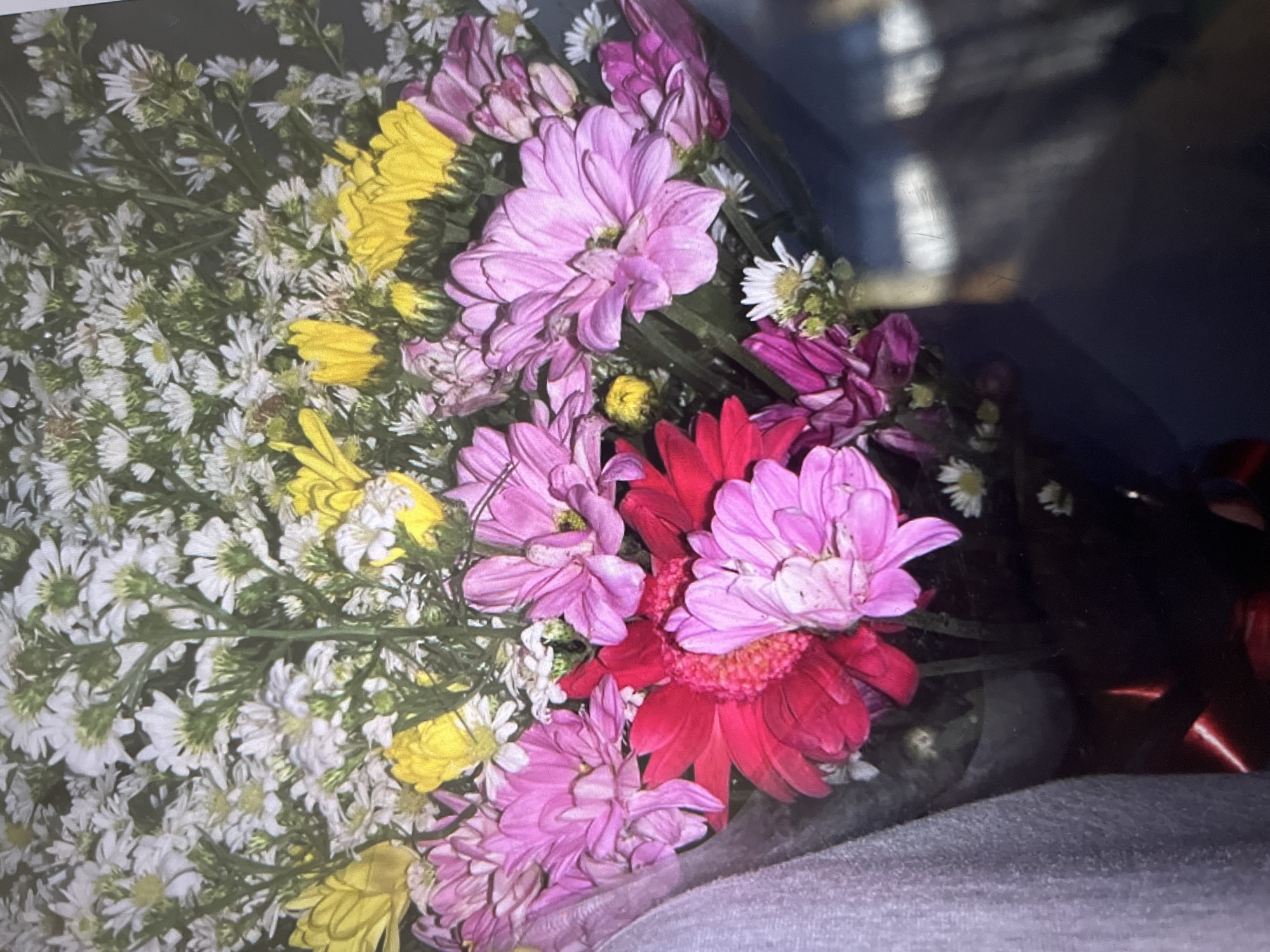 The image shows a close-up of a bouquet of flowers, predominantly chrysanthemums and asters. The bouquet features a variety of colors, including vibrant pinks, deep reds, bright yellows, and white blossoms. Some of the flowers are in full bloom, while others appear to be buds. The background is dark and out of focus, with some reflections suggesting an indoor setting or a dark surface. A textured, light-colored fabric is visible on the left side of the frame, partially obscuring the lower part of the bouquet. There are no people visible in the image. The scene does not provide any specific geographic markers to indicate a location in the city of Unknown, Unknown.