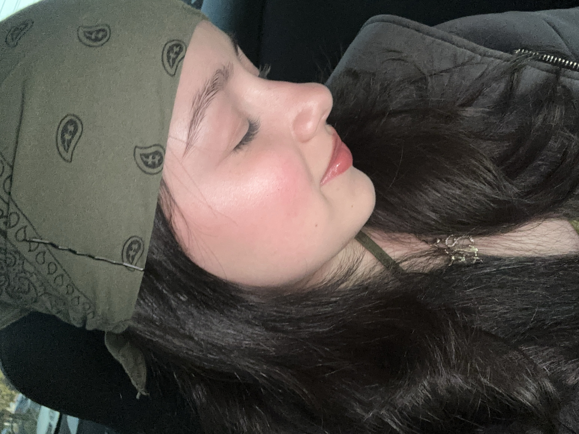 A young person with fair skin is seen in a close-up, profile-view indoor photograph. The person is lying down with their eyes closed, possibly sleeping. They are wearing an olive-green bandana with a paisley pattern on their head, and their dark, wavy hair is spread around them. The person appears to be wearing a dark-colored jacket or sweater and a light-green top underneath. A delicate necklace is visible around their neck. There are no visible indications of the location being Yerevan, Armenia.