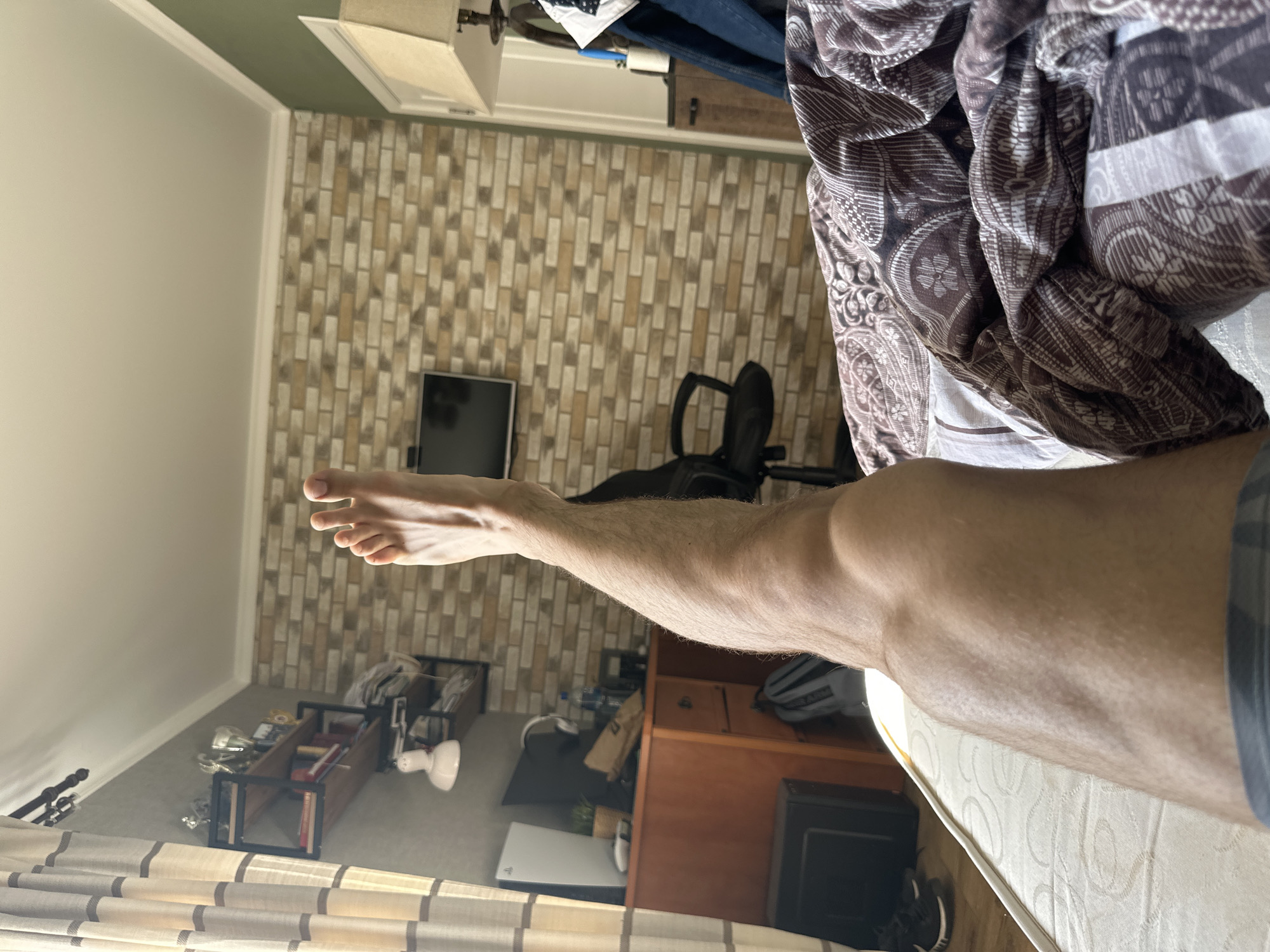 The image shows a room interior. Dominating the foreground is a lower leg and foot extending towards the upper left of the frame. The individual is lying on a bed with a patterned comforter. In the background, a patterned brick wall is visible, alongside a desk, chair, and TV. A bookshelf with various items is located in the lower-left corner. A window with curtains is also visible at the bottom of the frame. The location in the city of Unknown, Unknown is not provided.