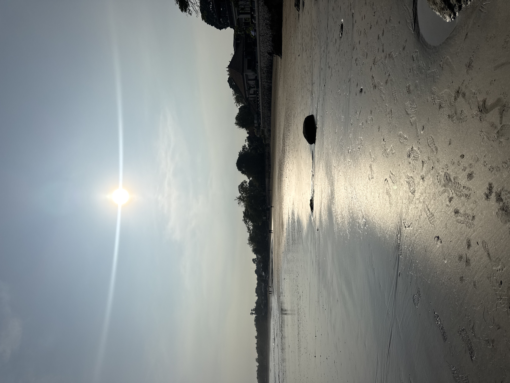 The image depicts a sunlit beach at Jimbaran, Indonesia. The sun shines brightly in a hazy sky. The beach is damp and reflects the sunlight. Footprints and various other marks are visible in the sand. A dark rock sits on the beach, its reflection visible in the wet sand. Buildings and trees line the edge of the beach.