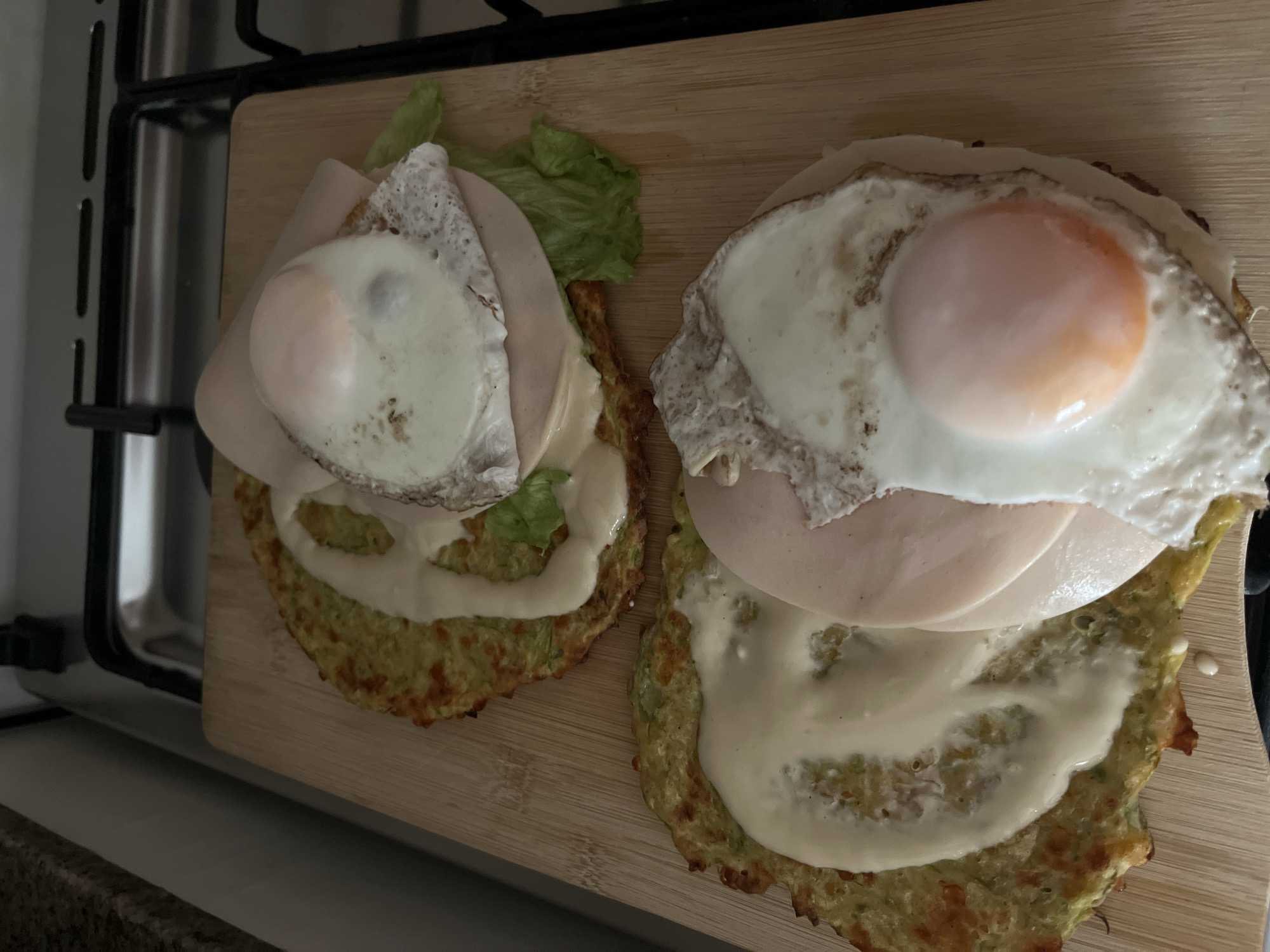 Two zucchini pancakes are topped with mayonnaise, ham, lettuce, and a fried egg. They are placed on a wooden cutting board, which is on a kitchen counter next to a gas stove. The lighting suggests it is daytime. There are no people or animals present in the image. The location is a kitchen.