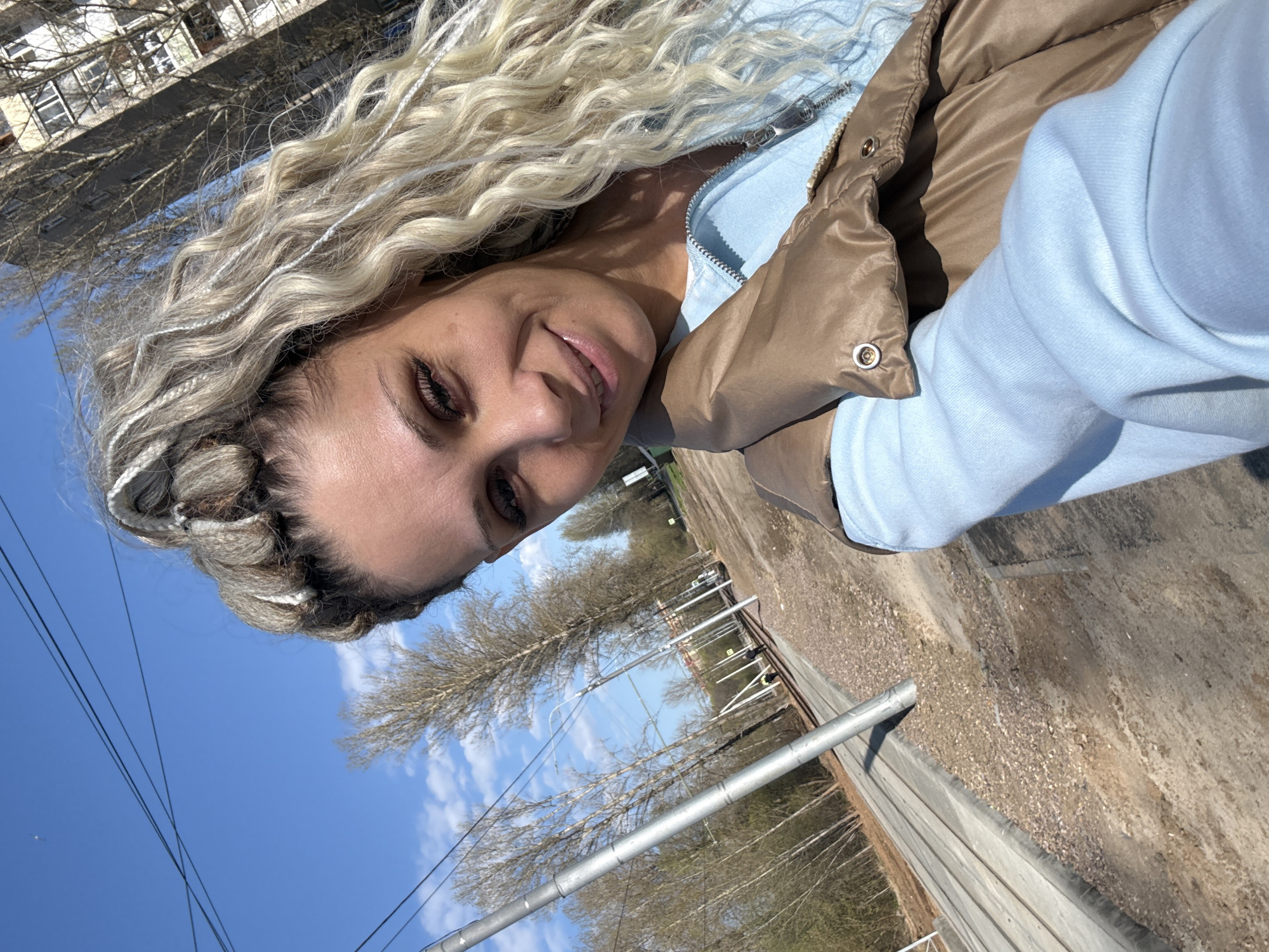 A woman with braided, light-colored hair is the central subject of the image. She wears a light blue hooded sweatshirt and a brown quilted vest. The background shows elements of an urban environment, possibly the city of Rusanovo, Russia, as requested. Trees and a building are visible in the distance. A fence and metal structures are also present in the foreground. The sky is blue with some clouds.