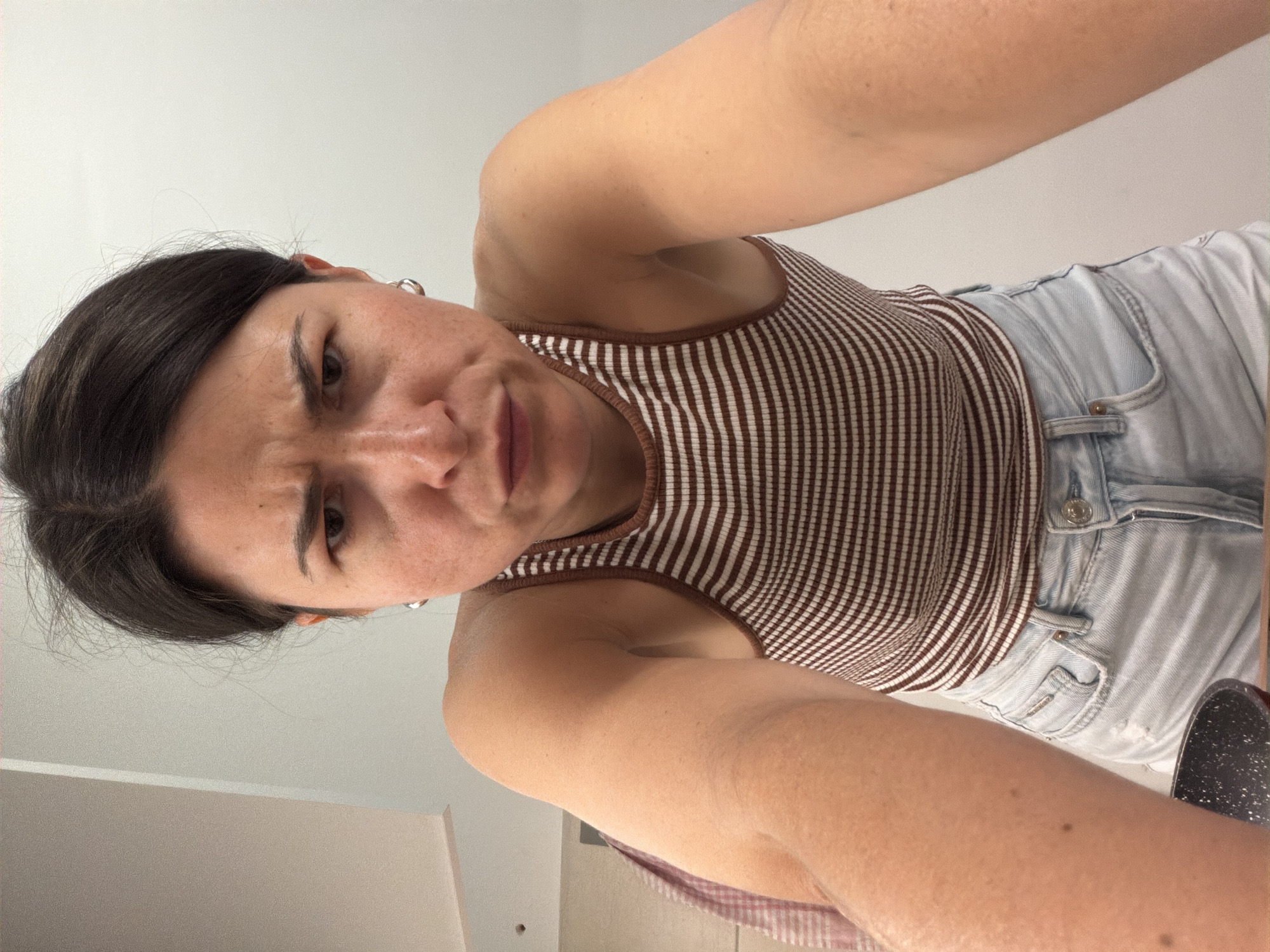 A woman with dark hair is angled from above, wearing a brown and white striped tank top and light wash jeans. Her expression is frowning. The background appears to be an indoor space with white walls. It is impossible to identify the location as the city of Budva, Montenegro.