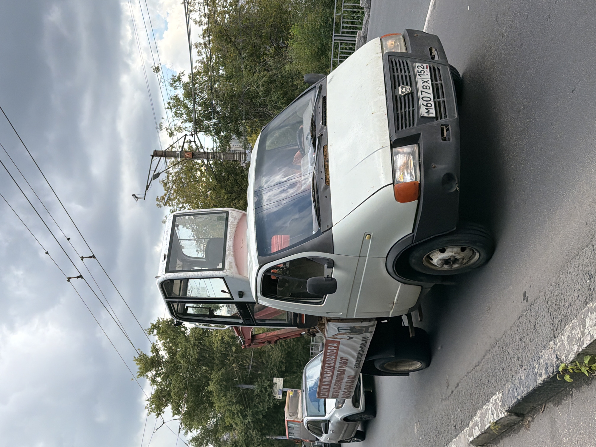 A white truck with a raised cabin and a small excavator attachment is driving on a paved road in Nizhny Novgorod, Russia. The truck has a banner on its side that reads "SERVICES MINI EXCAVATOR ALL TYPES OF EARTHWORKS" in Russian. The truck has a license plate that reads "M607BX 152 RUS". A silver car is driving behind the truck. Trees and power lines are visible in the background. The sky is cloudy.