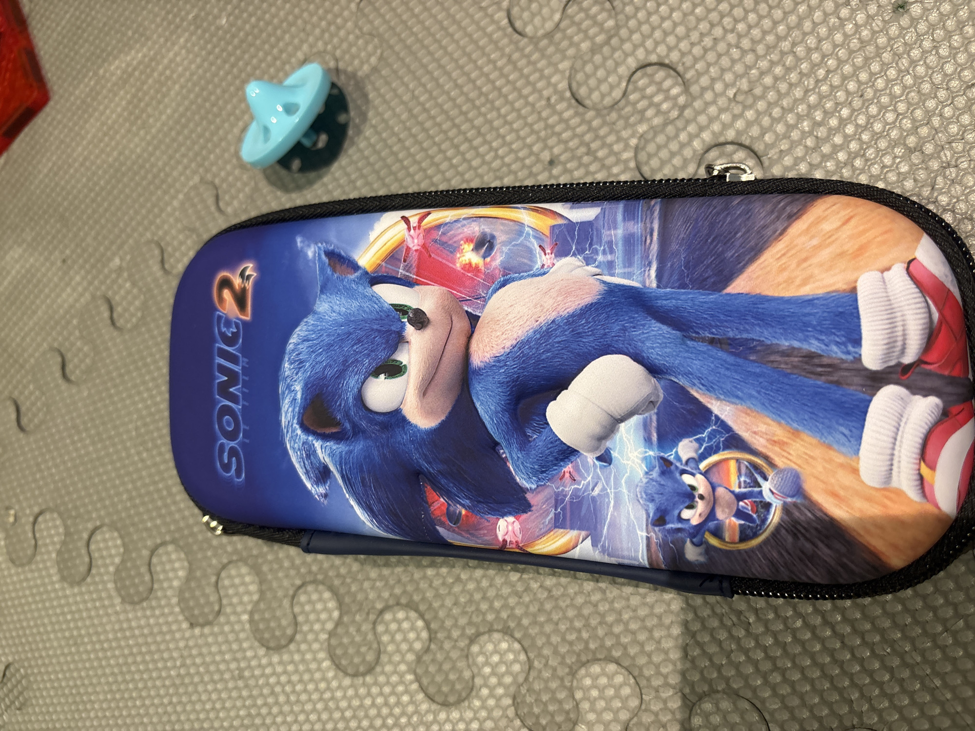The image shows a blue pencil case with the Sonic the Hedgehog 2 movie logo and characters printed on it. Sonic is depicted in a running pose, with a smaller version of himself in a ring behind him. The pencil case is lying on a textured grey surface, possibly a play mat or floor. To the right of the pencil case is a small, light blue spinning top toy. The background is slightly out of focus, but it appears to be an indoor setting. There are no discernable landmarks or clues that would indicate the location as Dubai, United Arab Emirates.
