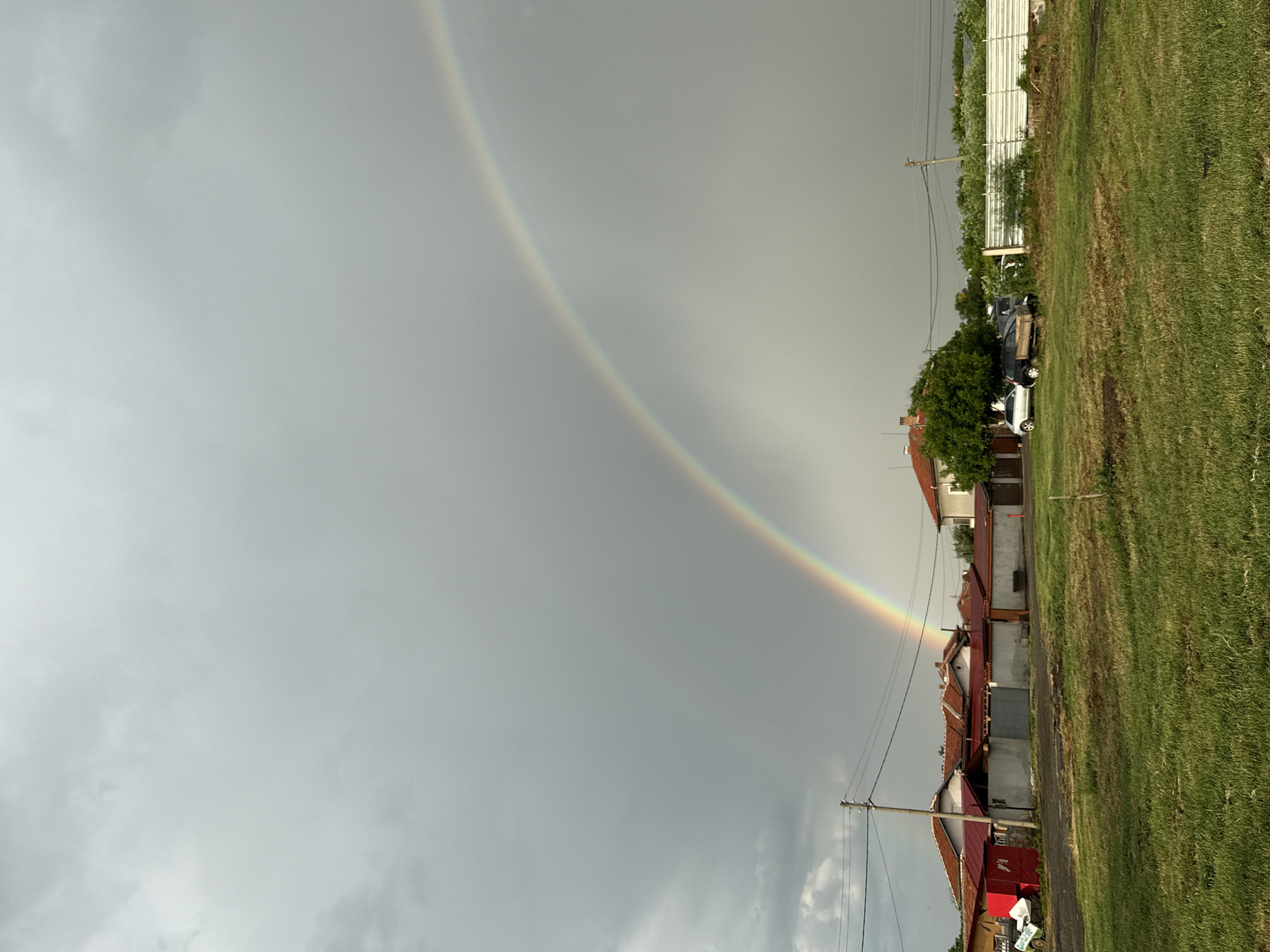 The image captures a rainbow arching across a gray, overcast sky in what appears to be a rural or suburban area of Sarnevo, Bulgaria. The rainbow's end appears to touch down near a cluster of modest homes with red-tiled roofs, suggesting its proximity to a residential area. A grassy field occupies the foreground, contrasting with the buildings. The photograph is taken at ground level. Utility poles line the edge of the residential area, indicating infrastructure development. The environment suggests recent rainfall or ongoing humid conditions. The absence of people and immediate action enhances the still, natural scene.