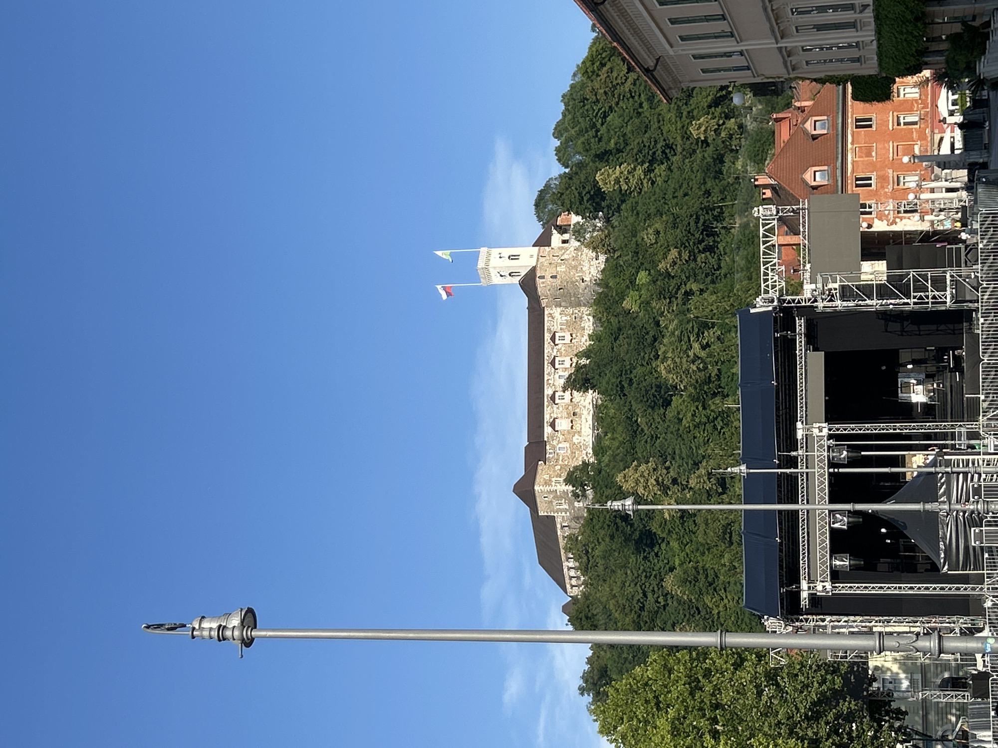 The image captures a wide shot of a city square with a castle on a hill in the background. In the foreground, a stage with a black backdrop and lighting equipment is set up. Several lampposts are visible throughout the scene. The castle, constructed of stone, is perched atop a hill covered in lush green trees. Two flags fly from a tower on the castle. The sky is clear and blue. The overall scene suggests an outdoor event or preparation for one in a historic urban setting.