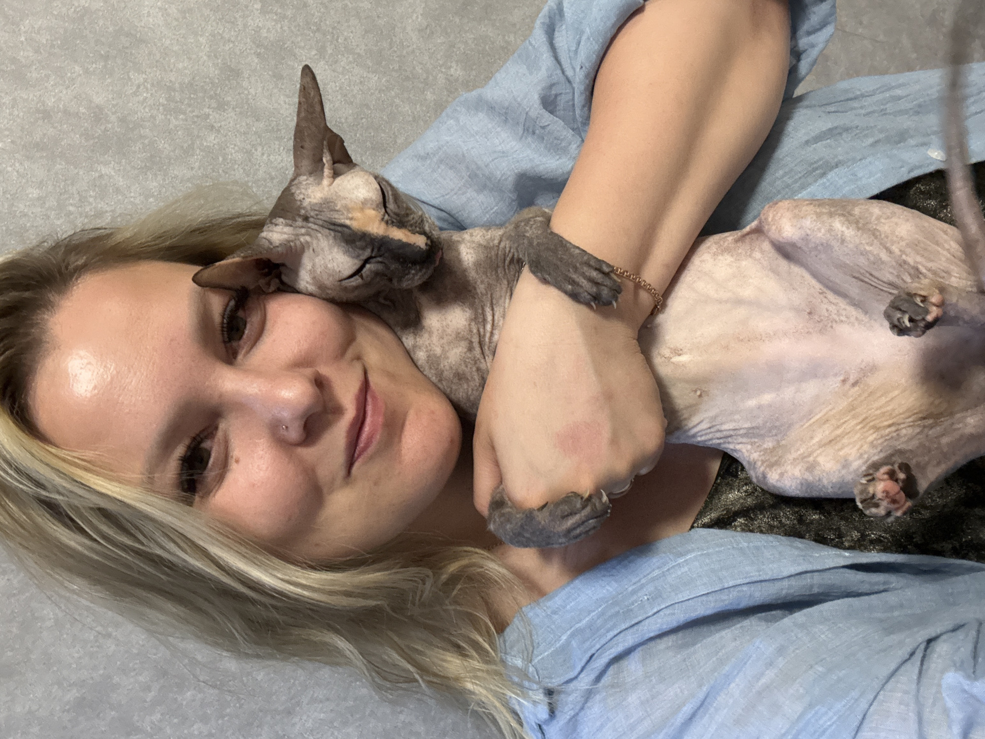 A woman with blonde hair and a nose piercing is lying down with a hairless cat on top of her. She is smiling and the cat is resting its head on her face. The woman is wearing a light blue shirt. The cat is lying on its back, with its paws in the air. The background is a gray surface. The location is unknown, but the city Марковский, Russia is specified.