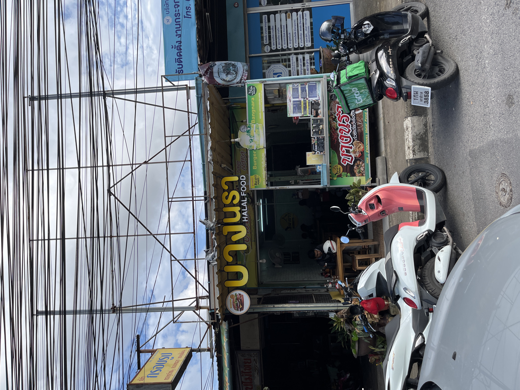 A street scene in Phuket City Municipality, Thailand, shows several shops and businesses lining a busy street. In the foreground, a grey car is parked, partially obscuring a white scooter and a pink and white scooter parked next to it. A black scooter with a green Grab delivery bag on the back is parked further down the street.
A halal food restaurant, "Bangnara Halal Food," is prominently displayed with a large yellow sign. A menu is visible on a stand outside the restaurant, and a man is seated at a table inside, looking at his phone. Another restaurant next door has a yellow sign with Thai script.
Overhead, a dense network of electrical wires crisscrosses the sky, characteristic of many urban areas in Thailand. The buildings appear to be constructed with concrete and metal, with awnings providing shade over the storefronts. The overall impression is one of a bustling commercial street in a tropical city.